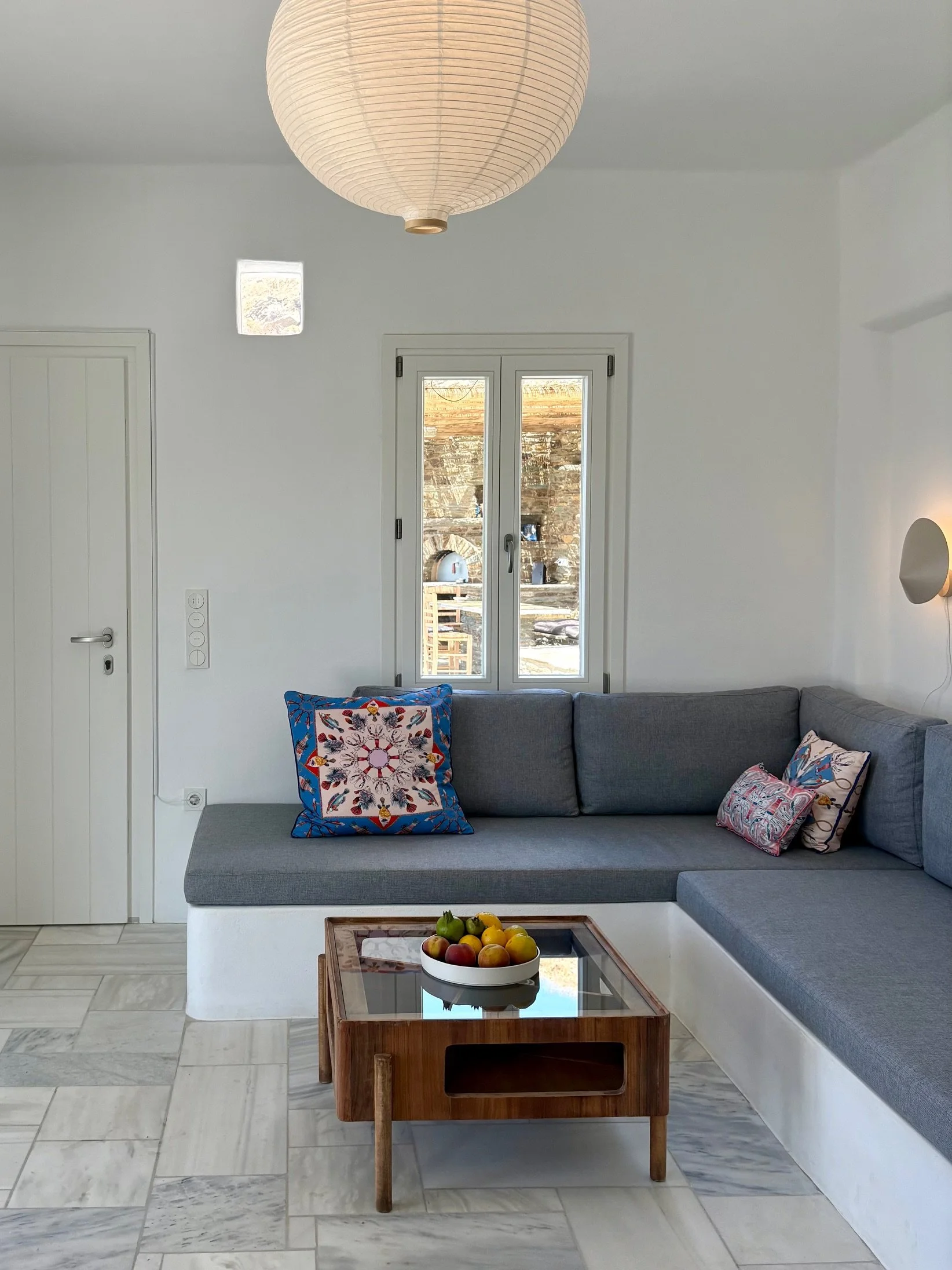 Living room with white walls, a gray sectional sofa with colorful throw pillows, a wooden coffee table with a bowl of fruit, a window showing an outdoor area, and a round paper lantern ceiling light.