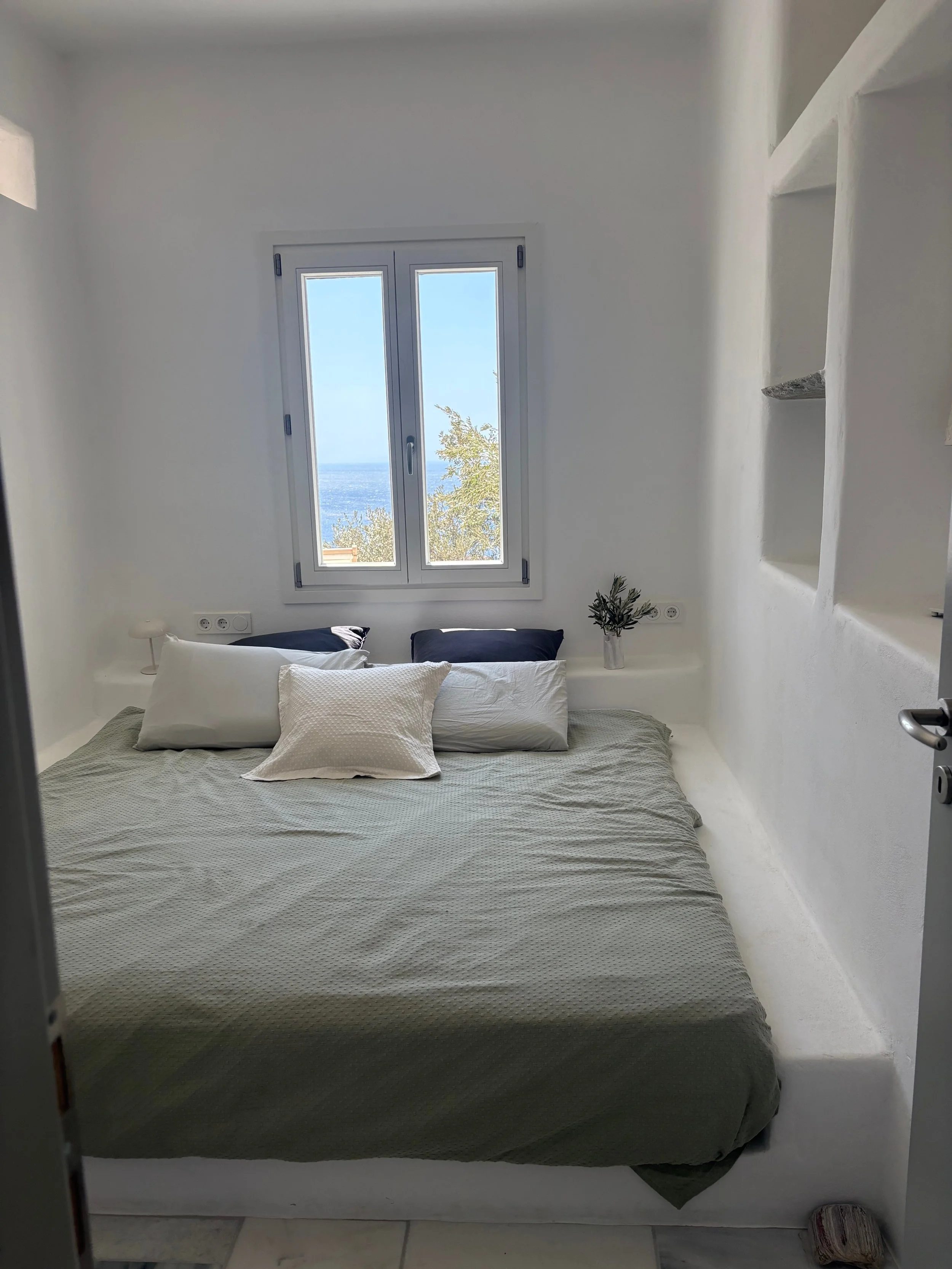 A minimalistic bedroom with white walls, a large bed with white and dark pillows, a window with a view of the sea, and built-in shelves on the right.
