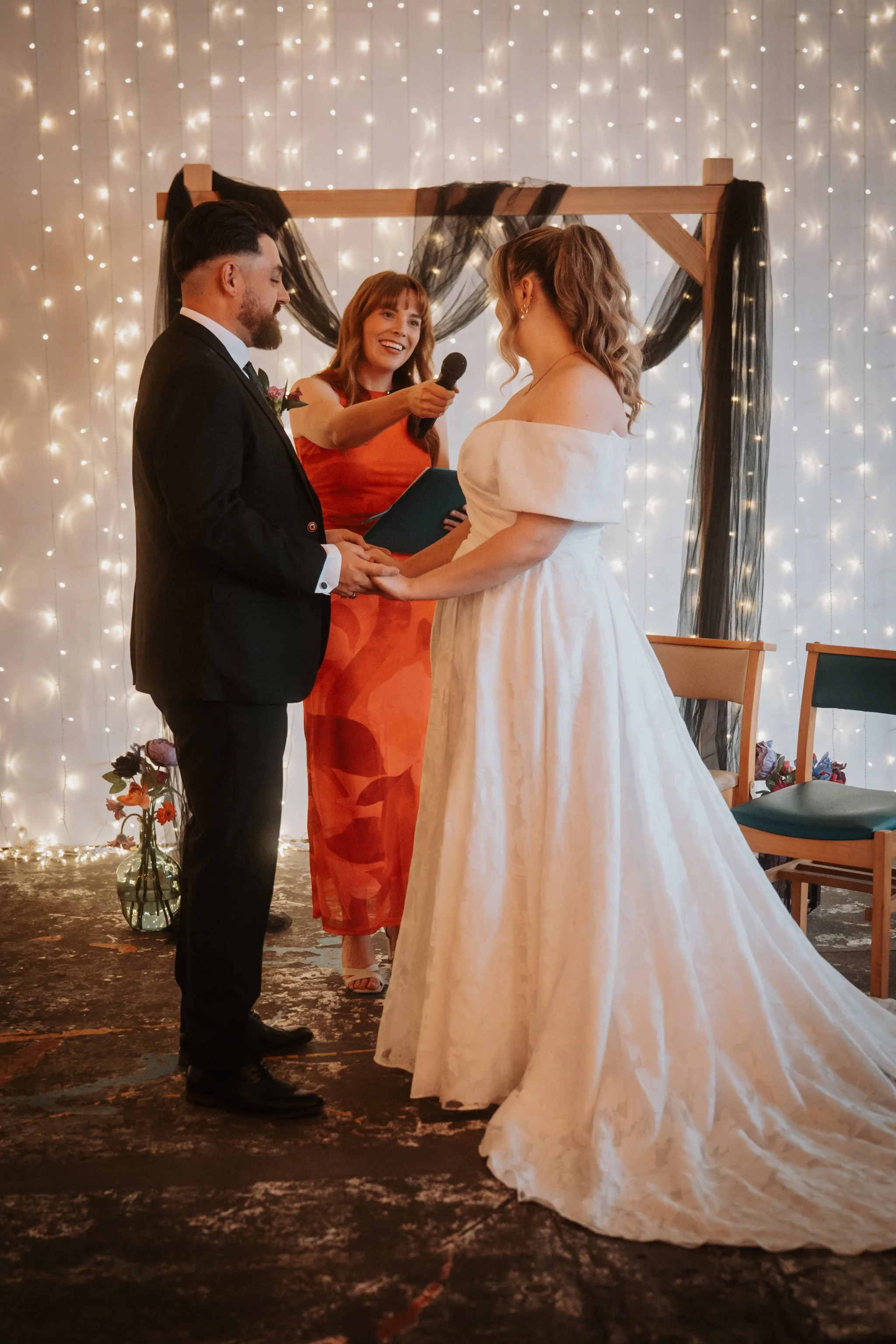A couple in wedding attire standing in front of an officiant during a wedding ceremony, with string lights and black decor in the background.