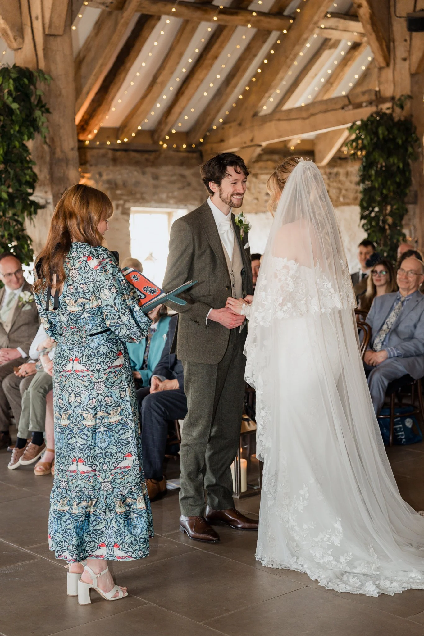 A wedding ceremony with a couple holding hands and facing each other, an officiant reading from a book, and guests seated in a rustic venue with wooden beams, string lights, and greenery.