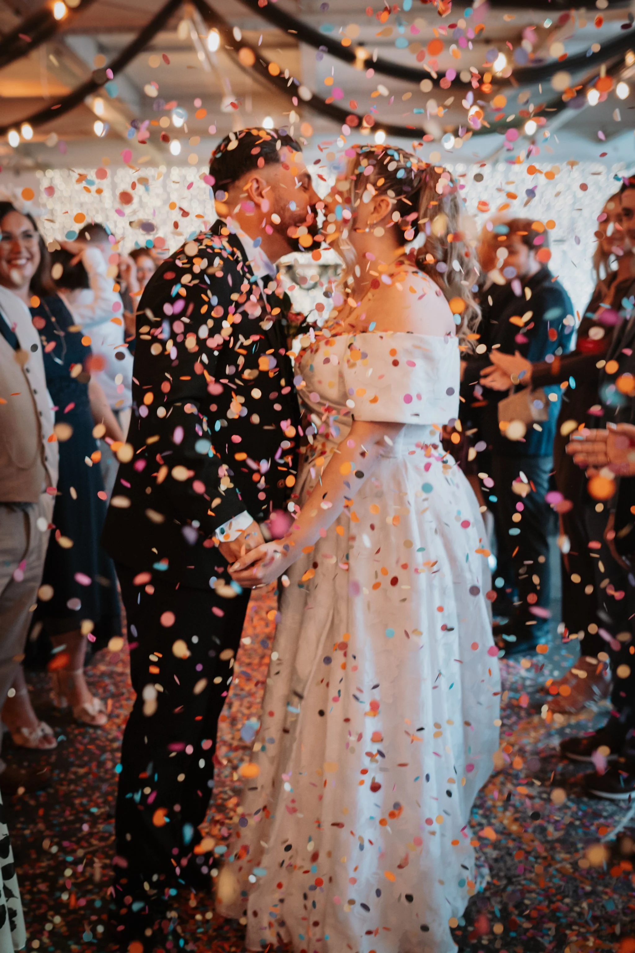 A couple getting married, holding hands and about to kiss, surrounded by colorful confetti at their wedding celebration, with guests in the background smiling and cheering.