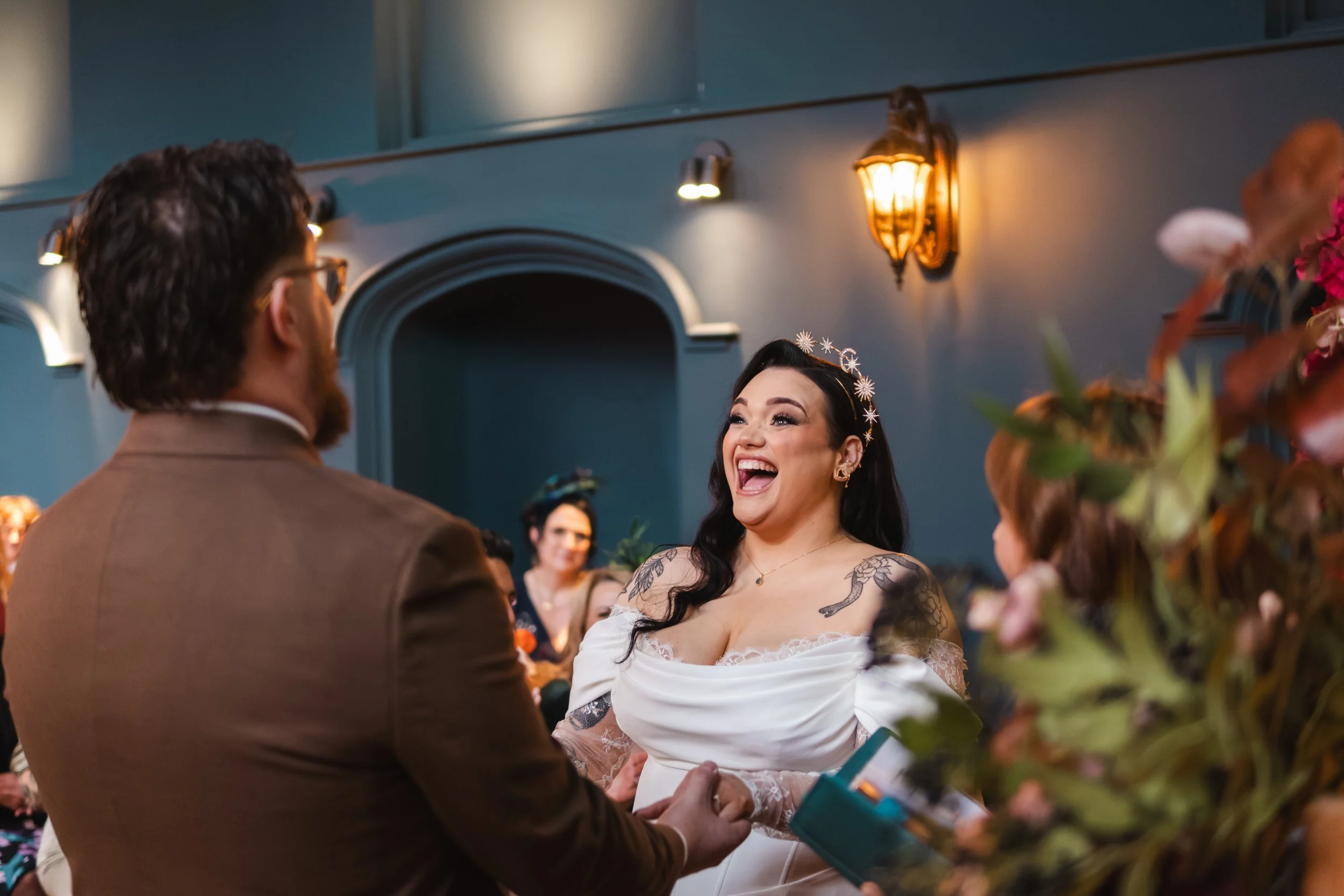 A woman with dark hair and tattoos on her shoulders, wearing a white off-the-shoulder dress and a flower crown, is smiling happily during a wedding ceremony. A man in a brown suit holds her hands, and other guests are visible in the background.