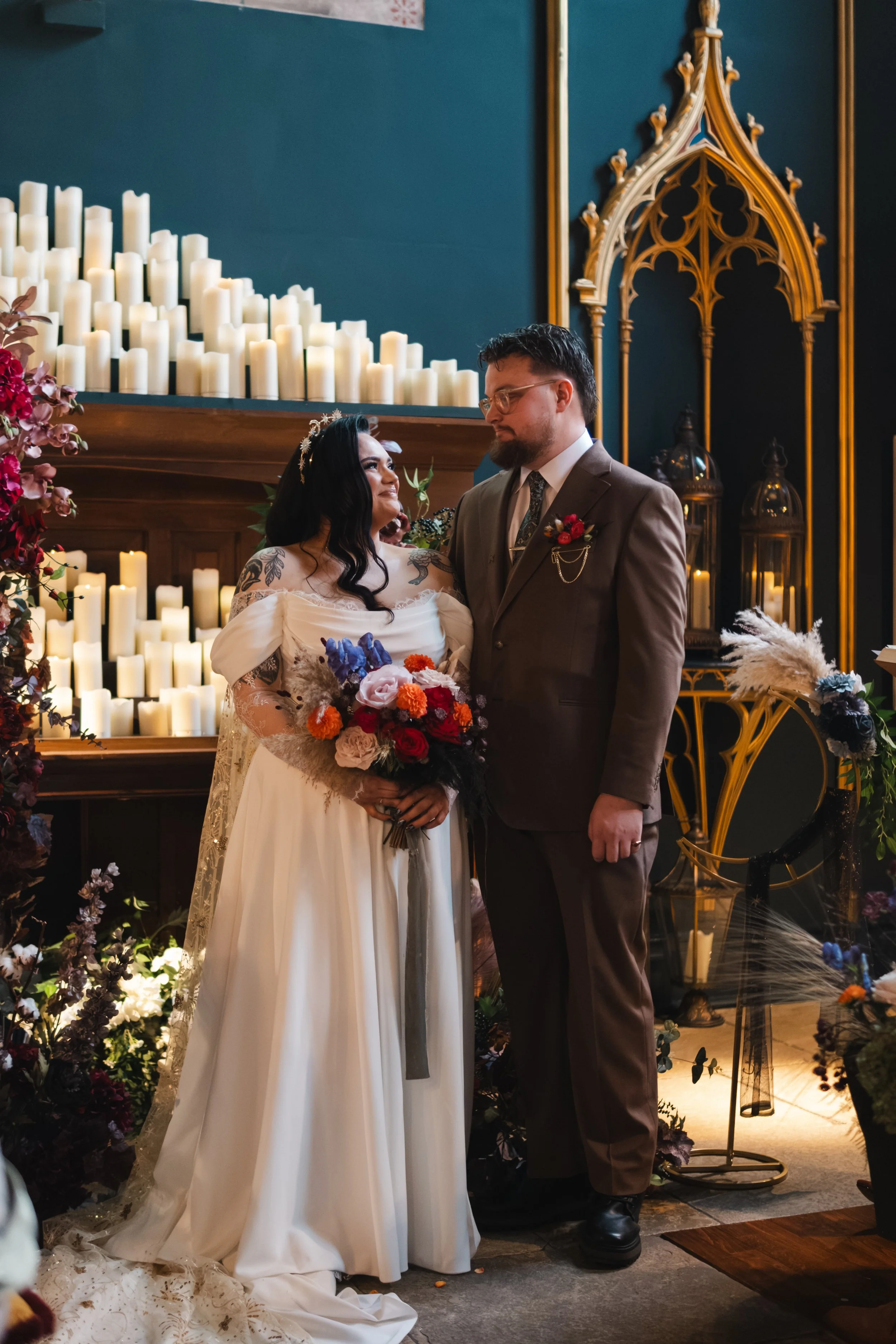A couple standing close during a wedding ceremony, surrounded by flowers and candles, with ornate gold decorations in the background.