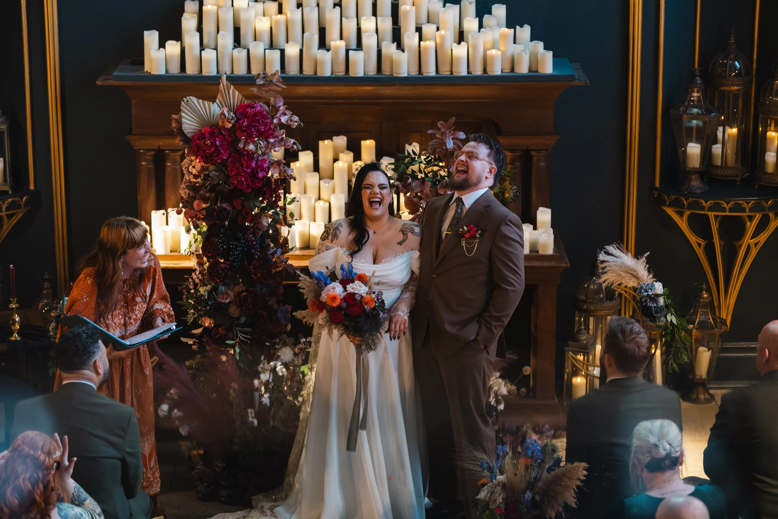 A wedding ceremony with a couple smiling and laughing, standing in front of a candlelit backdrop, surrounded by floral arrangements, with guests seated and an officiant on the side.