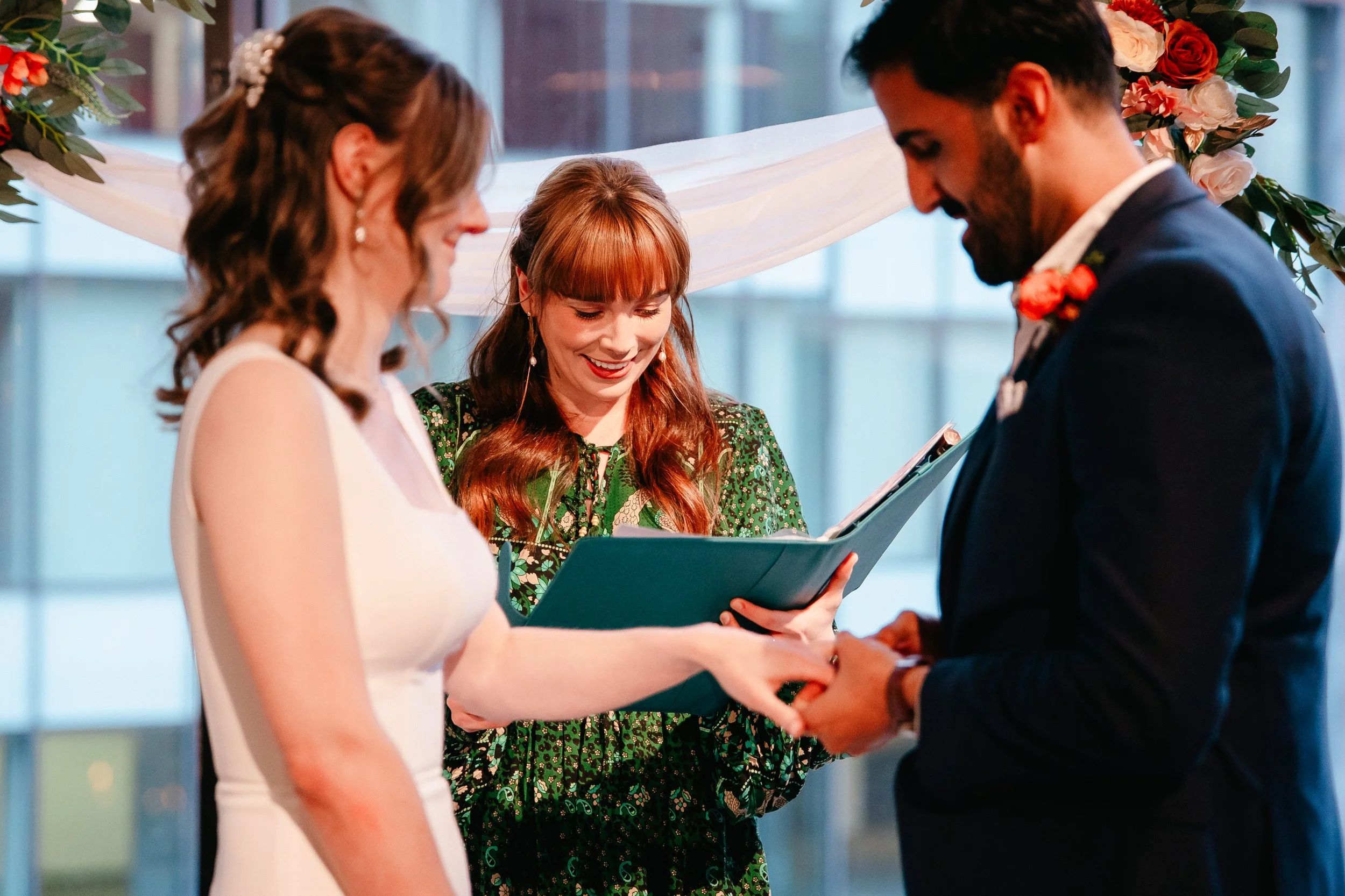 A couple getting married in an indoor ceremony, exchanging vows, with a officiant presenting a book or binder to them. The bride and groom are standing in front of a floral arch with greenery and white and pink flowers, and a large window in the background.