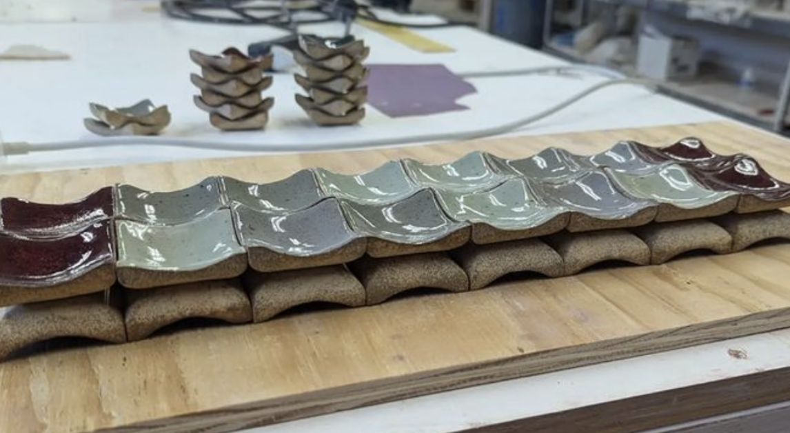 A long wooden board with clay tiles partially glazed in maroon, light green, and dark green colors, arranged in a pattern. In the background, there are stacks of unglazed clay tiles on a white table.