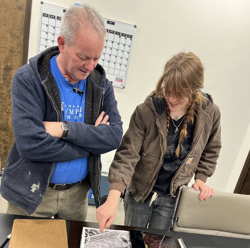 A man and a woman are standing at a table, looking at a black-and-white map, with the woman pointing at it. The man has short gray hair, wearing a blue shirt and a dark zip-up hoodie. There is a tablet and a notebook on the table in front of them, and a whiteboard calendar on the wall behind them.
