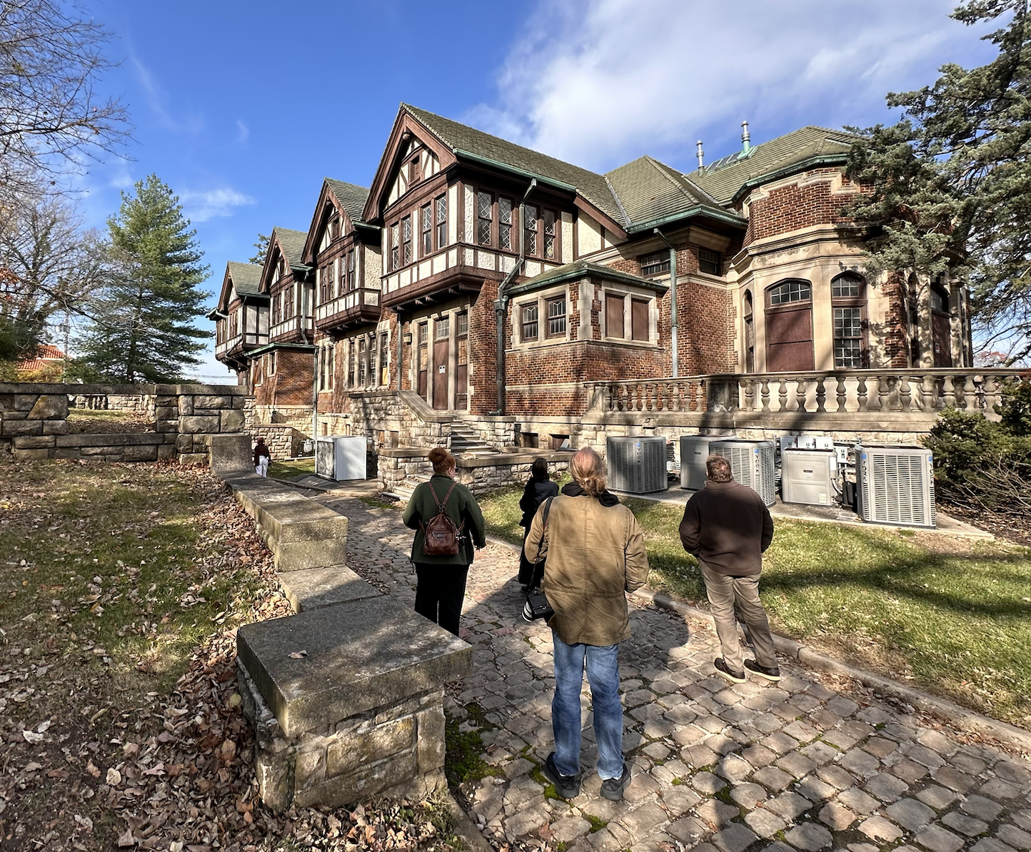 Group of people walking on a cobblestone path towards a large, historic brick house with multiple gables and a turret, under a partly cloudy sky.
