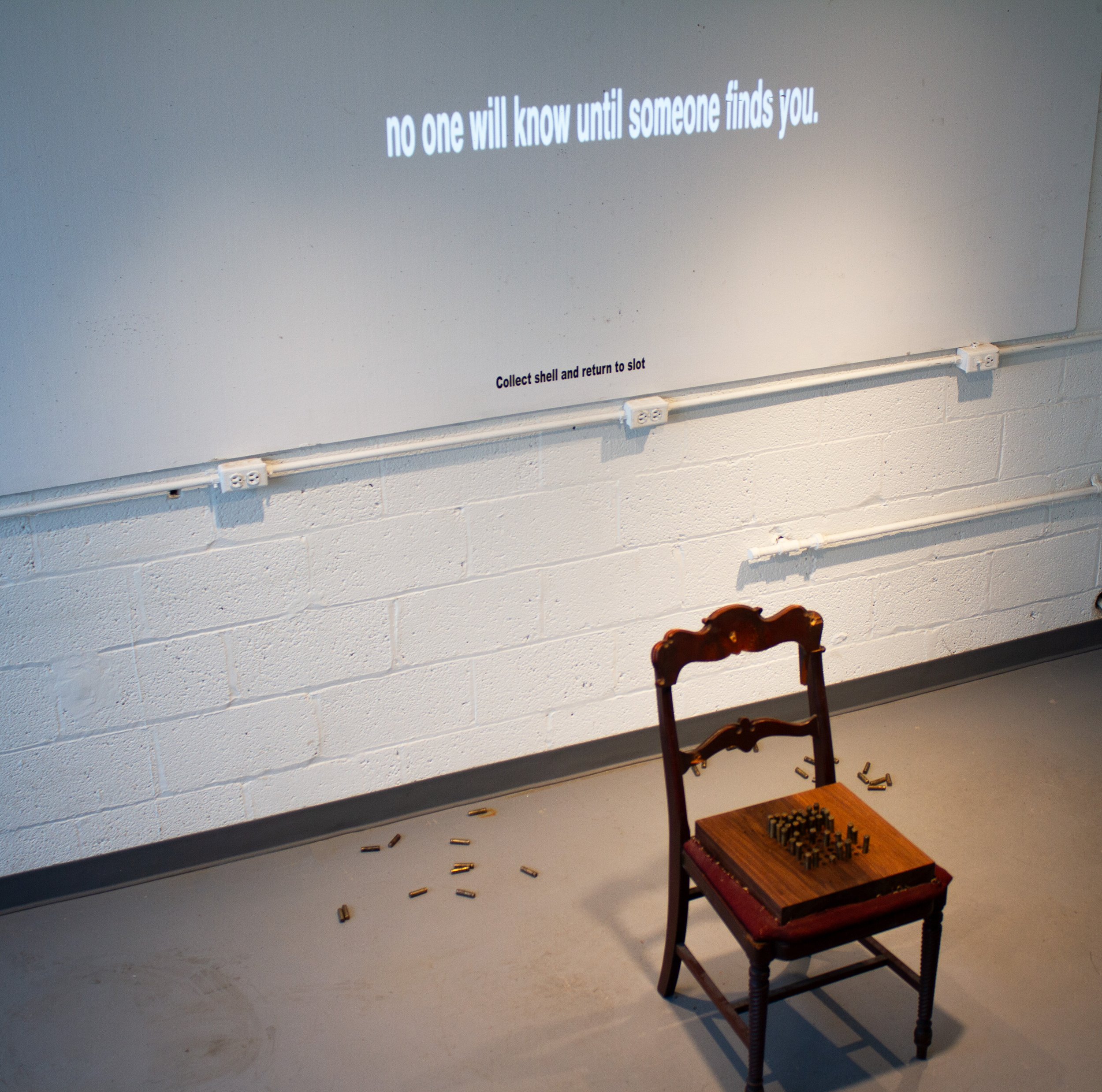 An art installation on a white brick wall features a large projection with the text 'no one will know until someone finds you' and instructions to collect a shell and return it to a slot. A vintage wooden chair with a chessboard on the seat is placed