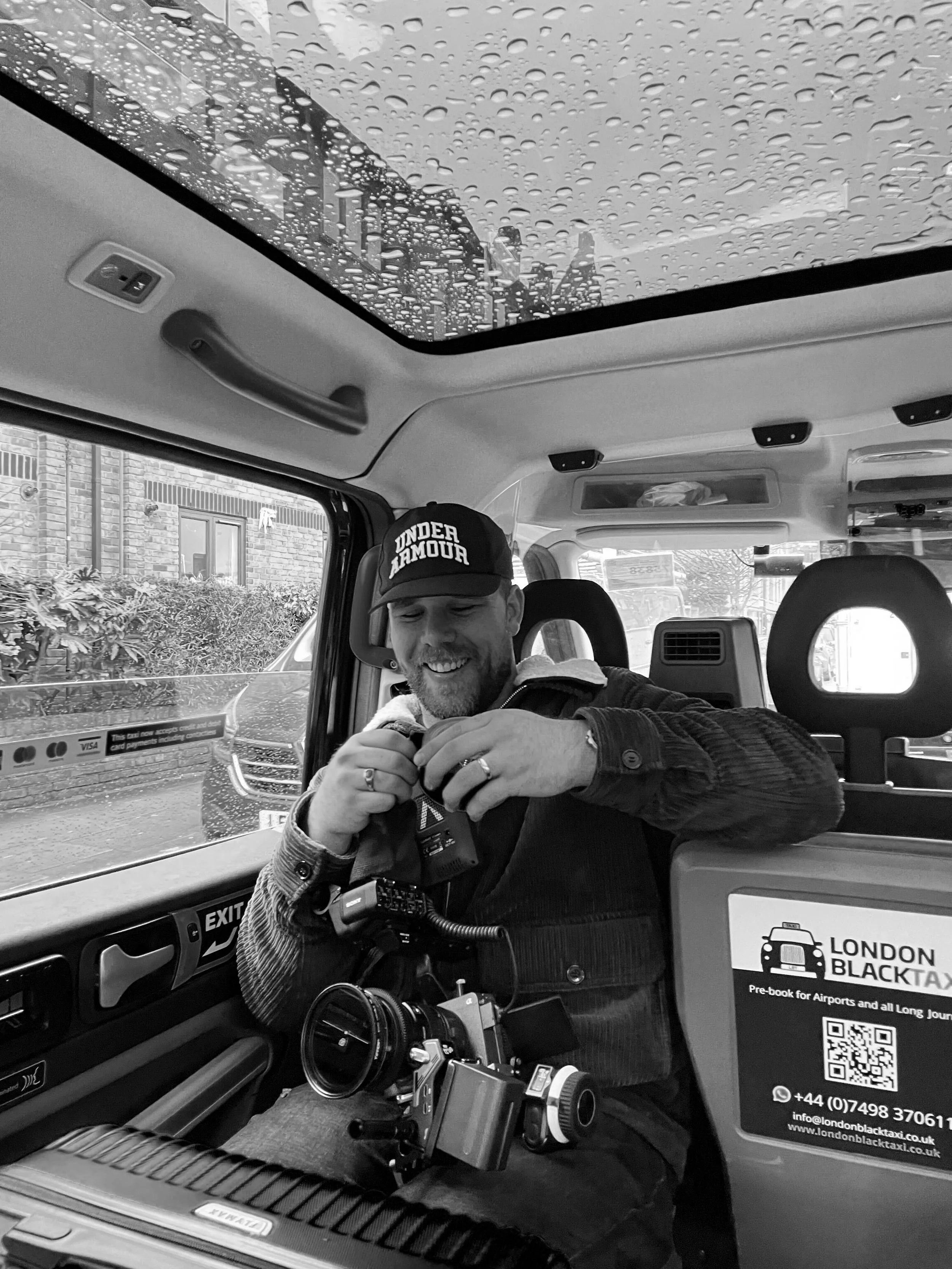 Man with camera gear inside a vehicle with raindrops on sunroof, wearing a cap, smiling, black and white photo.