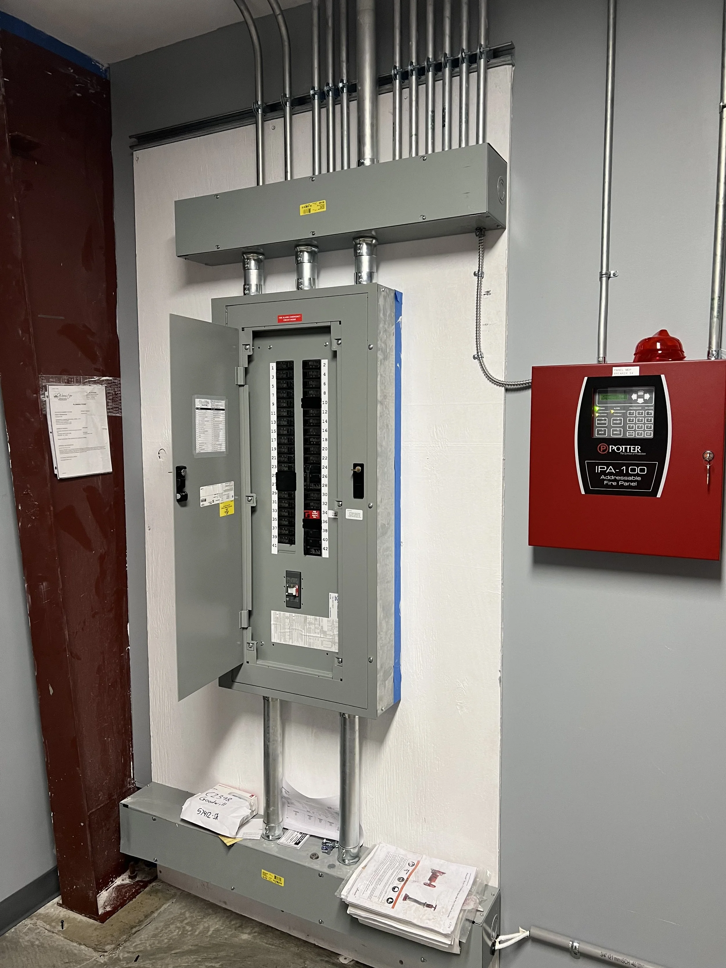 Electrical panel with open door, control box, and fire alarm panel on a wall.