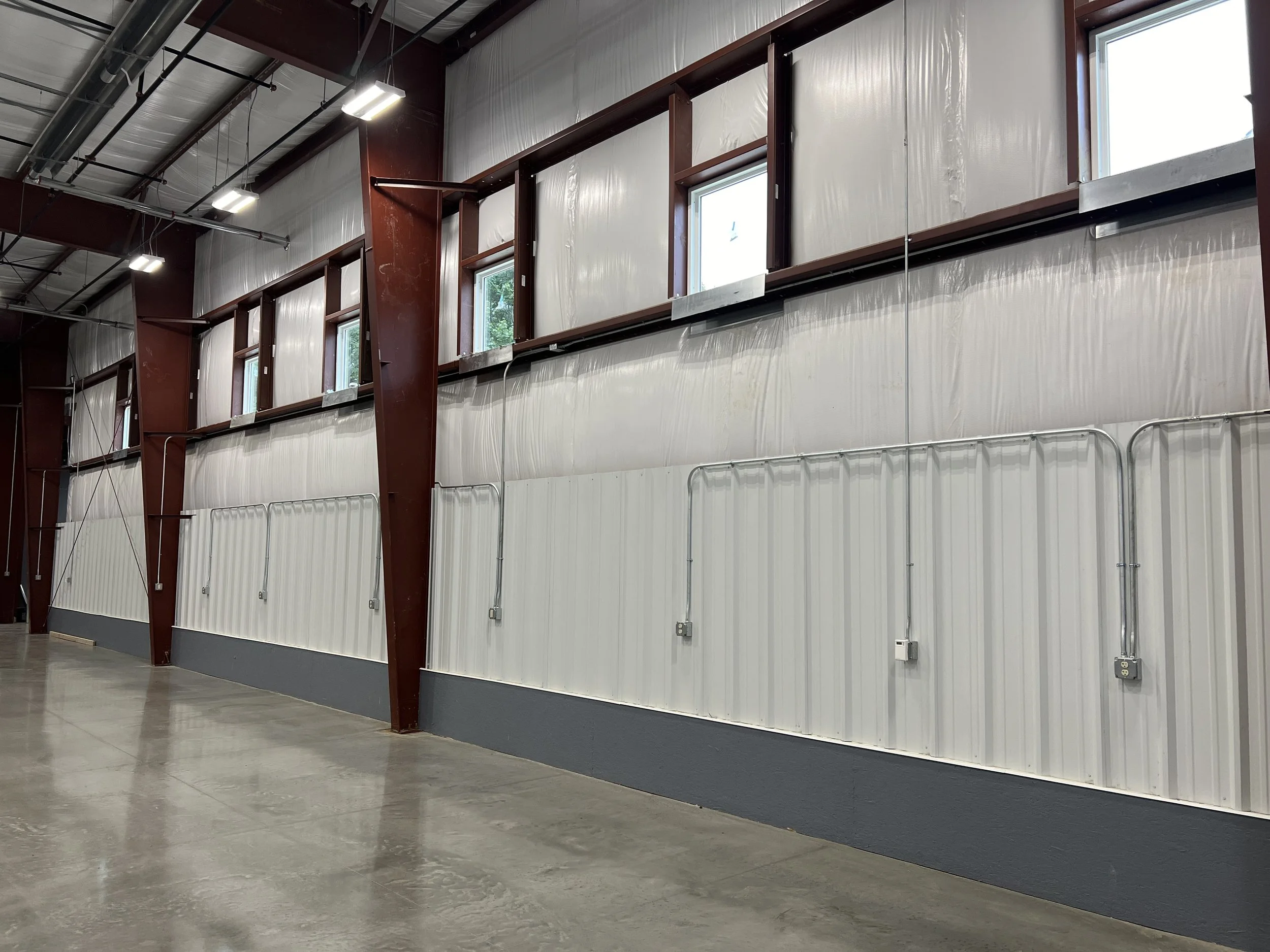 Interior of an industrial warehouse with metal walls, exposed beams, multiple small windows, and electrical conduit piping.