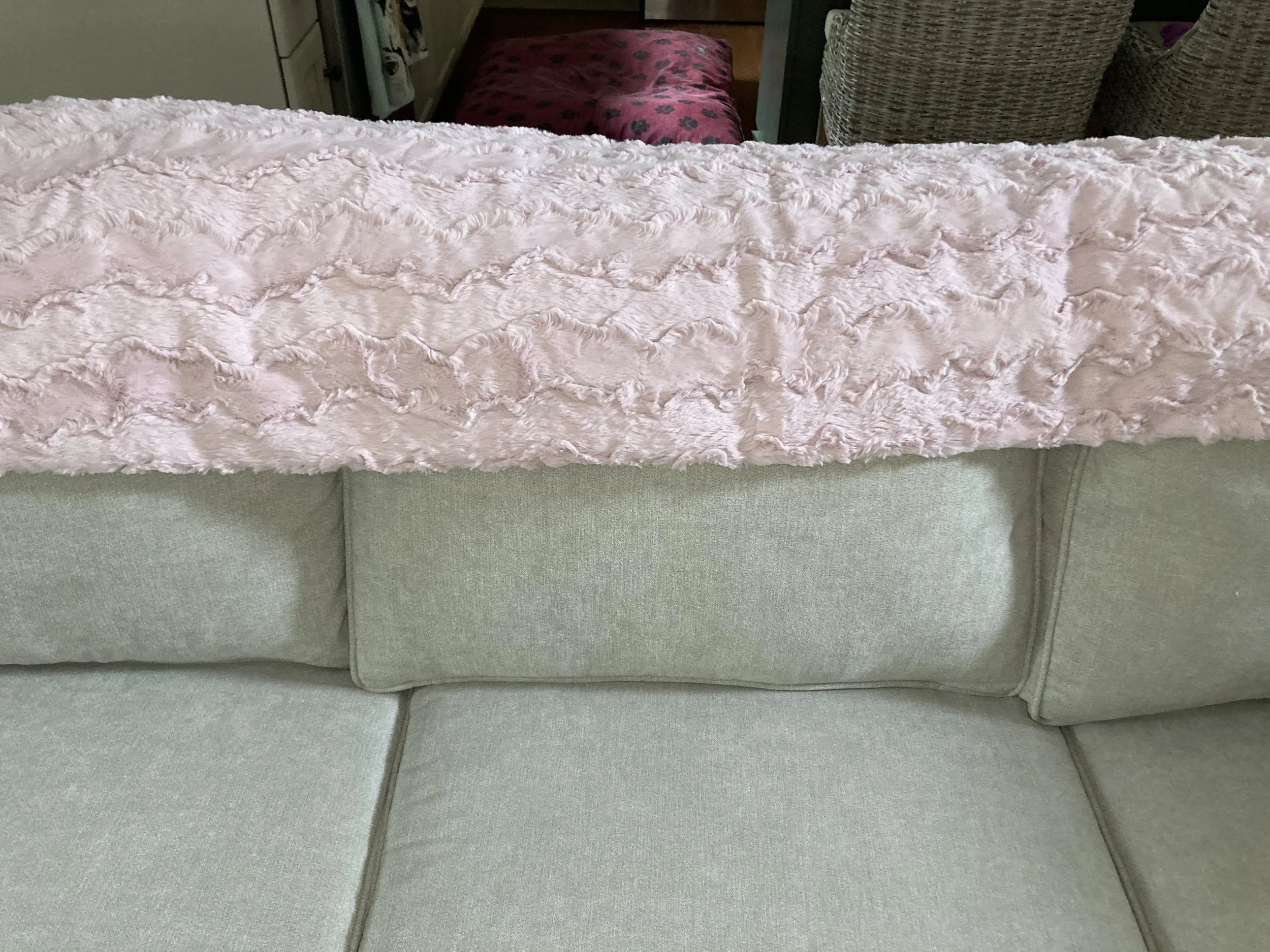 High-angle view of a plush Ice Pink Luxe Cuddle throw blanket draped over a neutral grey sectional sofa, showing the wavy embossed texture and soft pink hue.