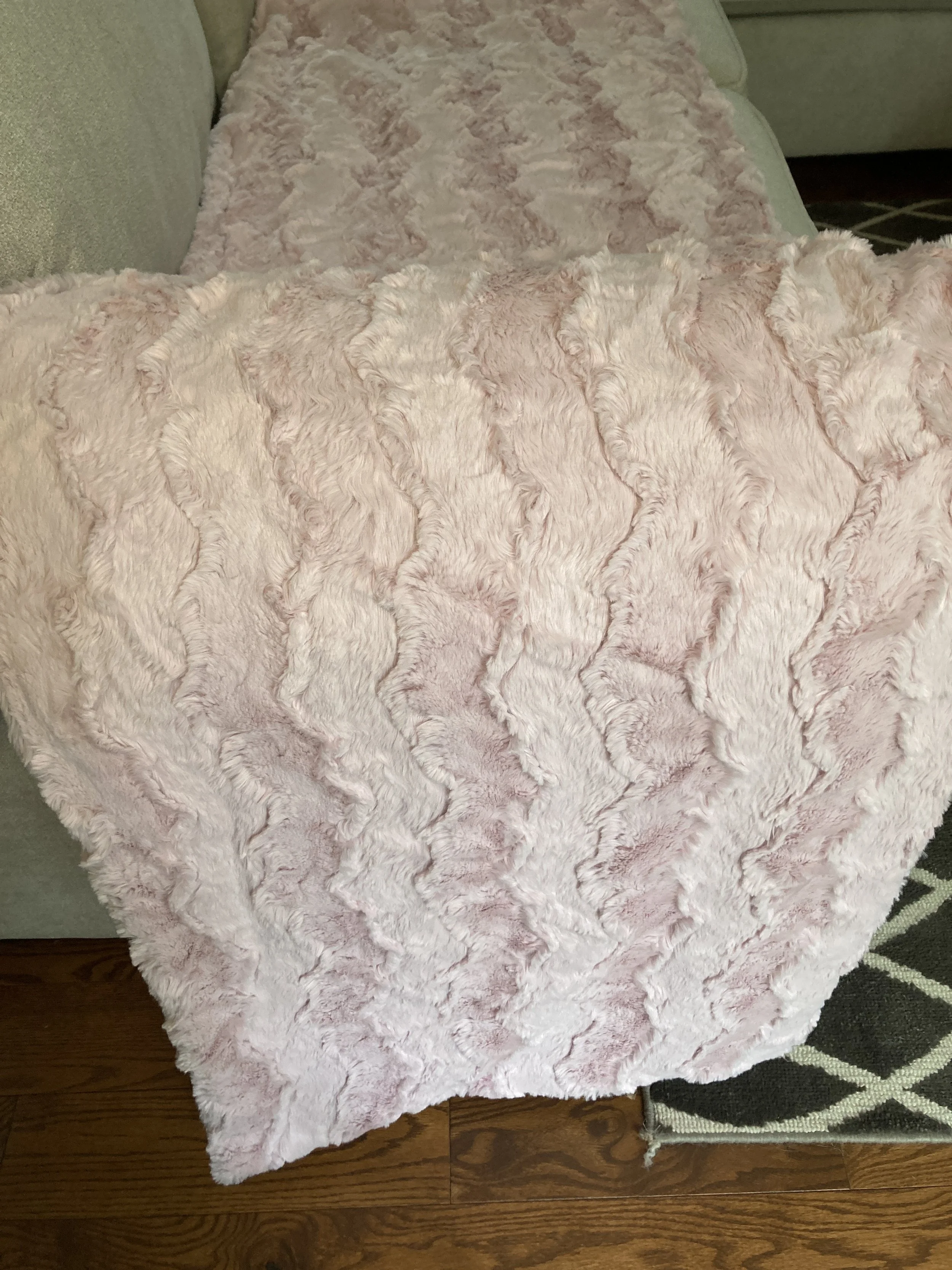 A double-sided pink faux fur blanket folded over the back of a grey couch, highlighting the thick, high-quality edges and luxurious drape of the Shannon Fabrics material.