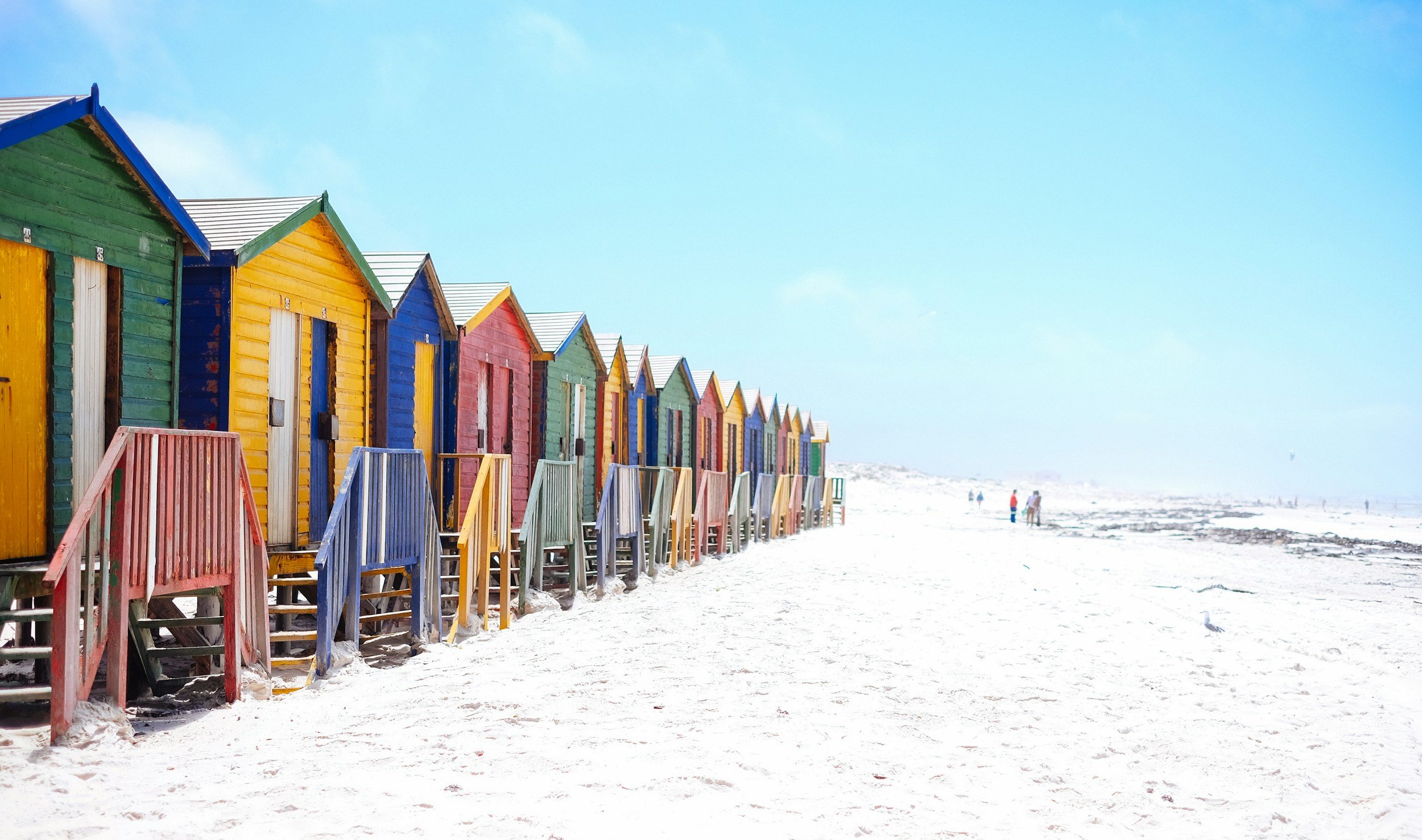 Colorful beach huts lined up on a sandy shoreline under a clear blue sky.
