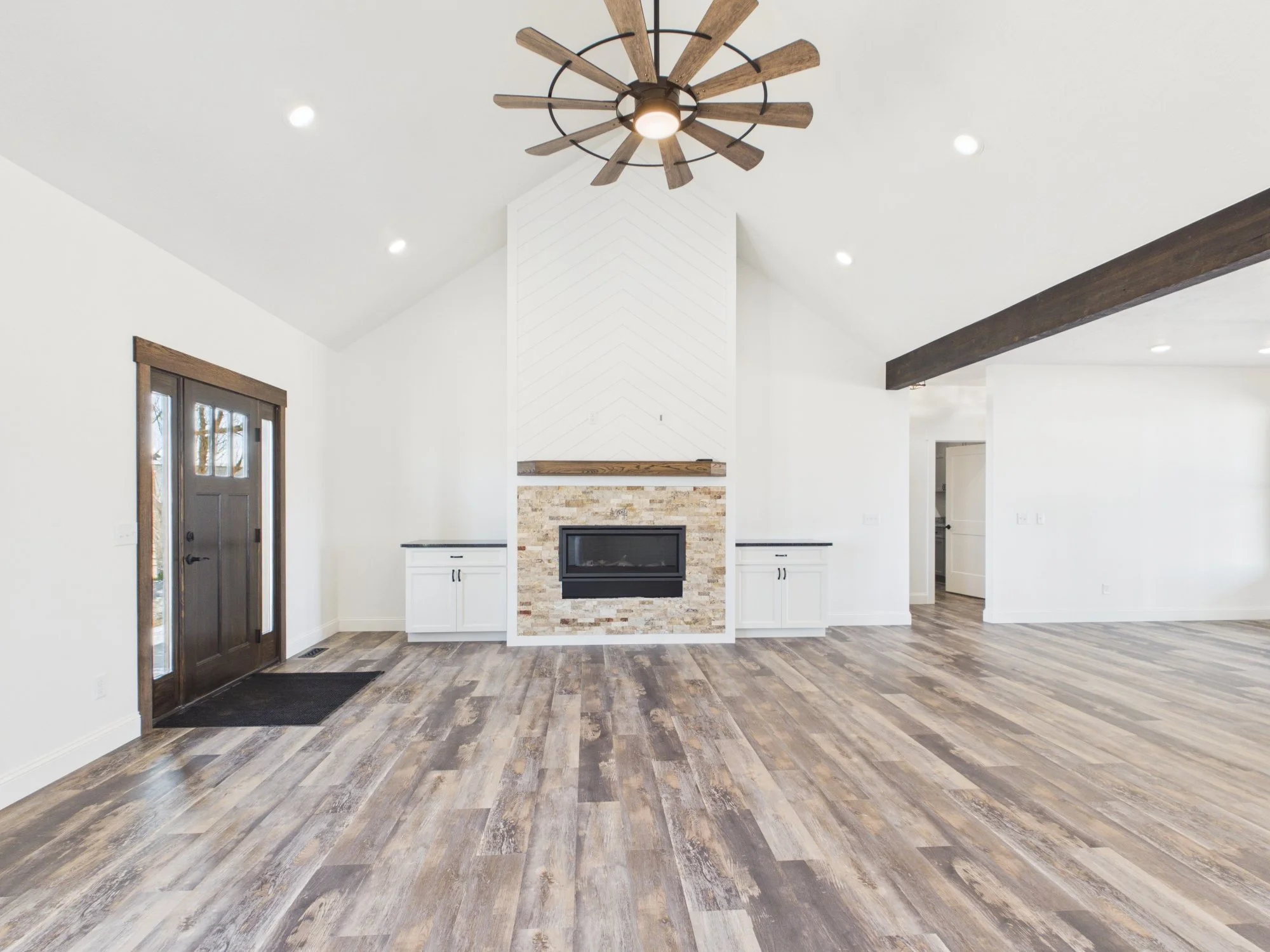Interior view of a spacious, modern living room with a white fireplace, wooden beam accents, a wood-look floor, and a ceiling fan with a wooden blade design.