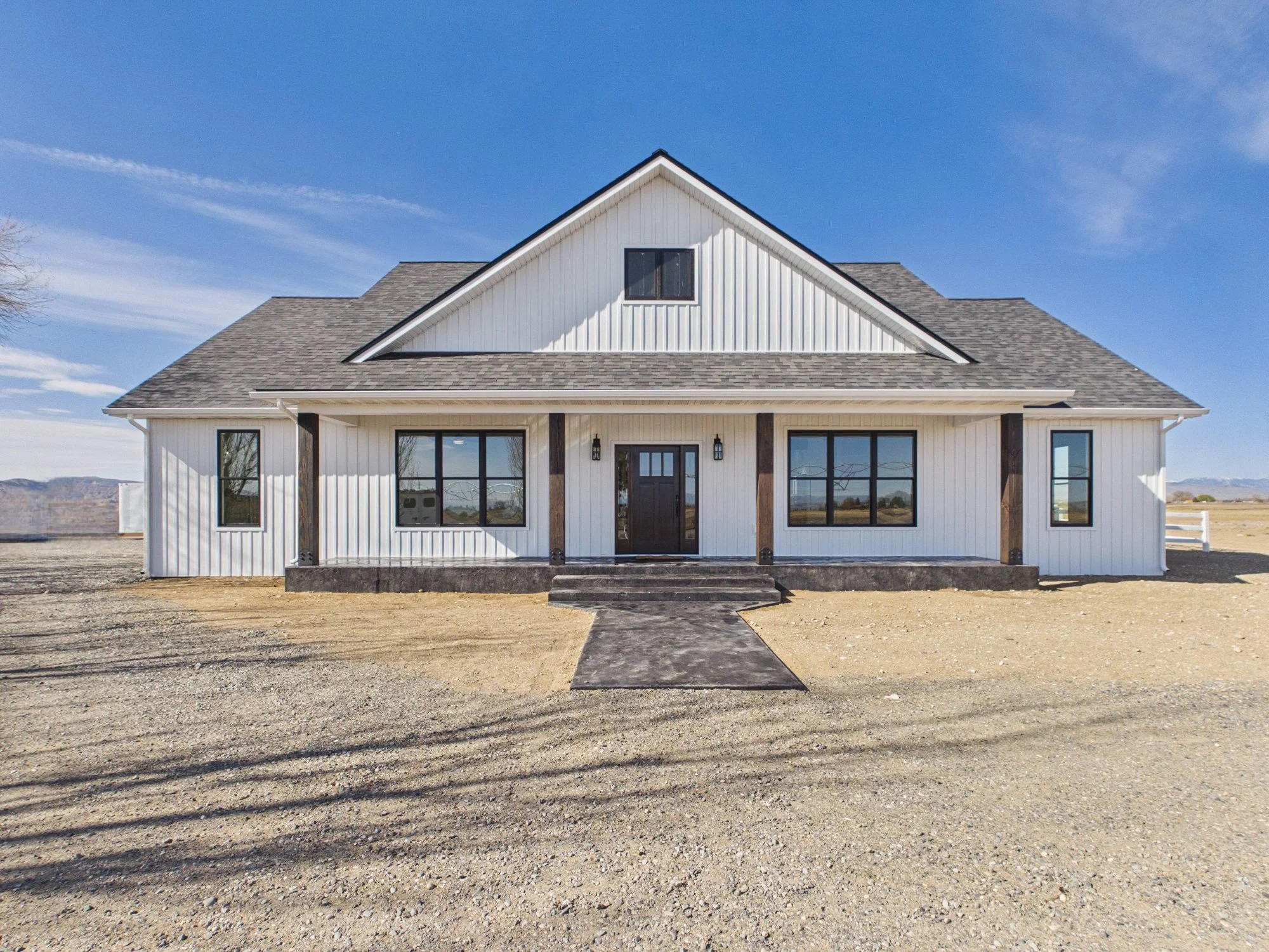 Front view of a modern white house with a gray roof, large windows, and a small porch with steps leading up to the front door. The house is situated in a barren landscape with minimal vegetation, under a clear blue sky.