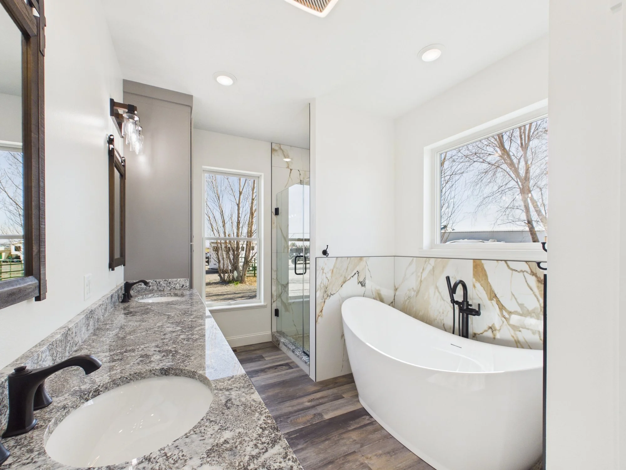 Modern bathroom with white walls and large windows, featuring a freestanding bathtub with black fixtures, a granite countertop with a sink, a marble accent wall behind the bathtub, and a glass shower enclosure.