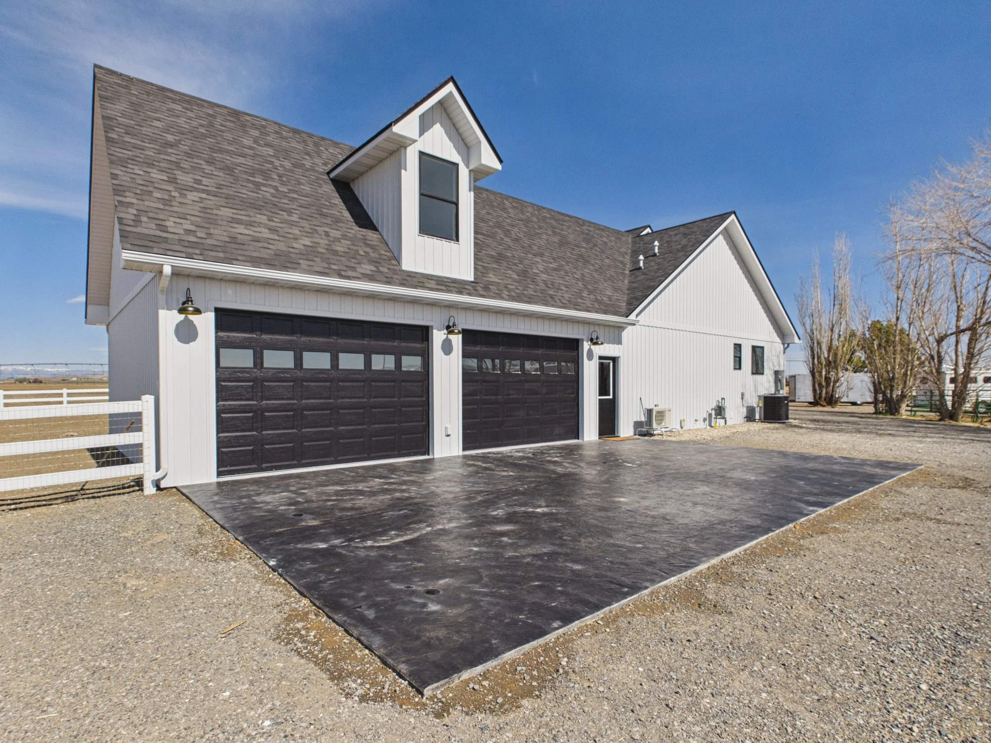 A modern white house with black garage doors, a large front porch, steep gable roof with shingles, and a dormer window. There is a gravel driveway and some leafless trees nearby under a clear blue sky.