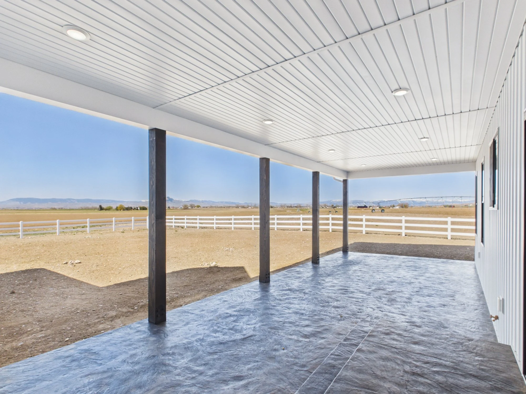 Covered patio area with a concrete floor and support beams, overlooking a rural landscape with open fields, fences, and mountains in the distance under a clear blue sky.