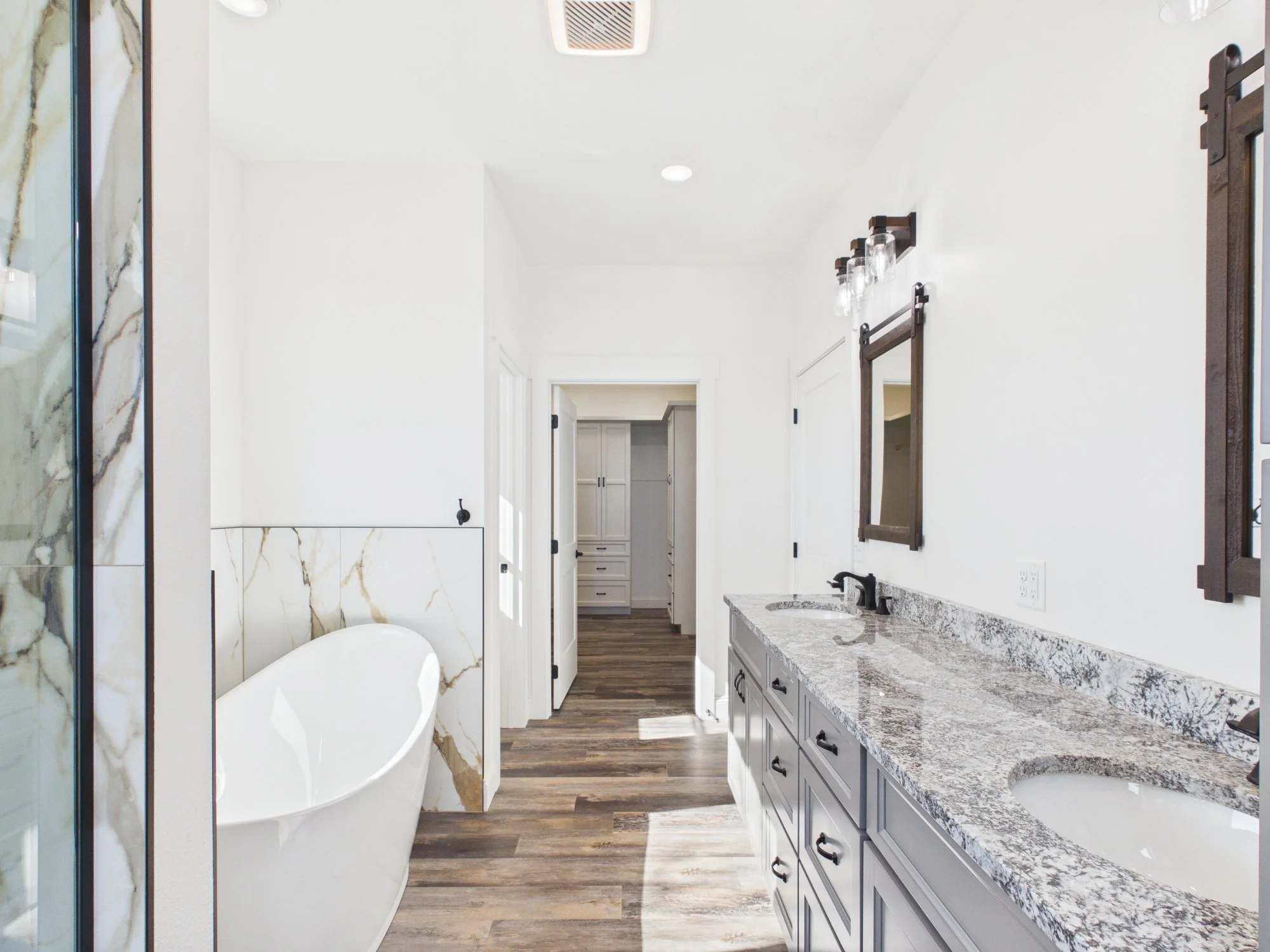 A modern bathroom with white walls, a marble shower wall, a bathtub, a granite countertop with a sink, a mirror and lighting, and a view into a closet area.