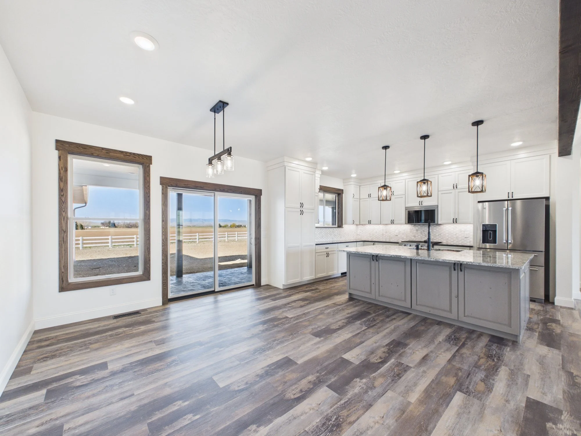 Interior view of a modern kitchen with white cabinets, dark hardware, hardwood flooring, pendant lights, and large windows showing an outdoor landscape.