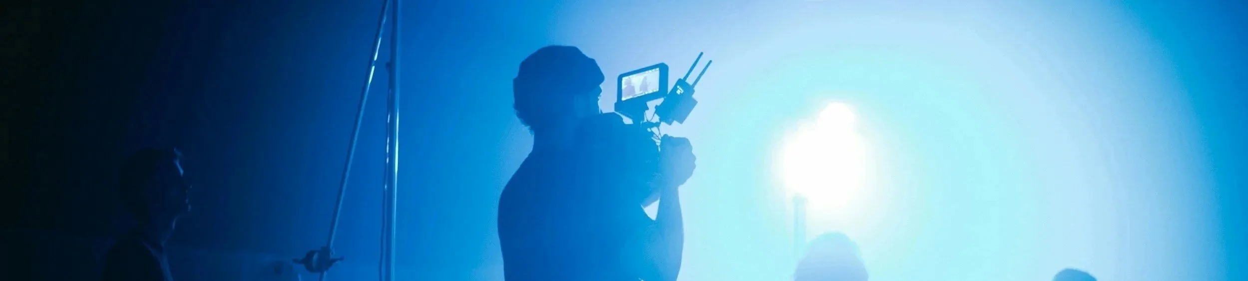 Silhouette of a person operating a camera rig in a blue-lit environment with fog or smoke, alongside other figures in the background.