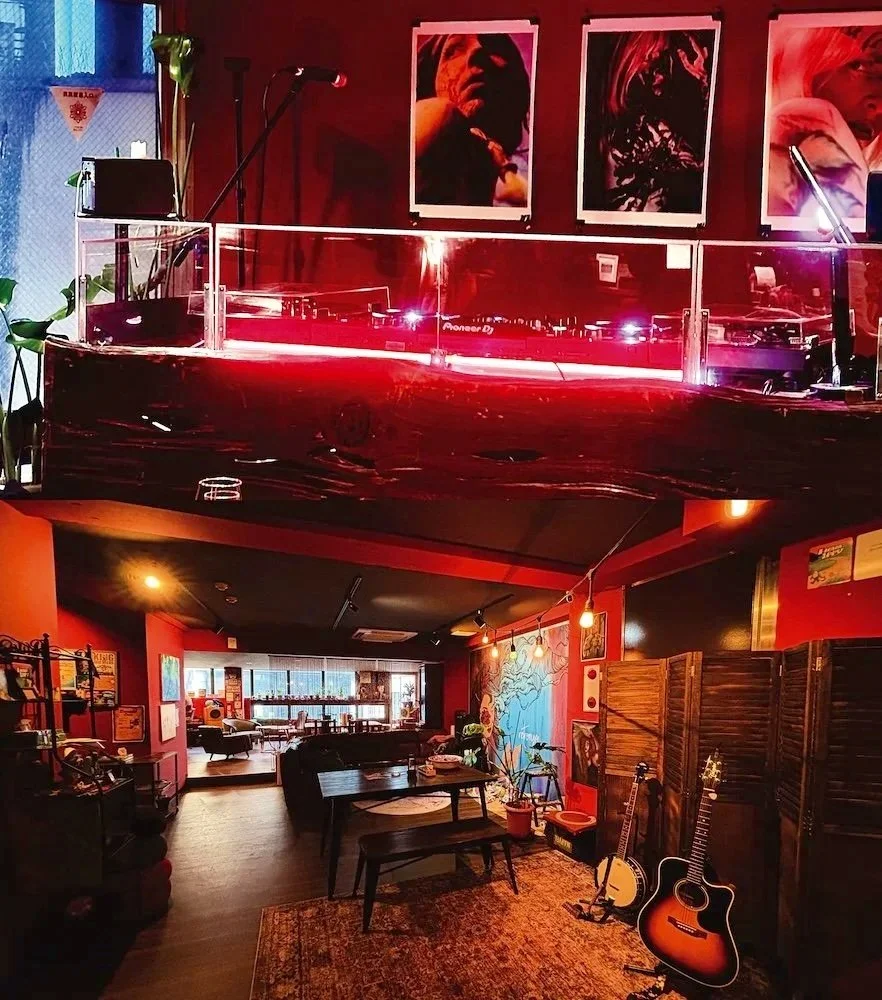 Interior of a cozy music venue or cafe featuring a jukebox, guitars, framed photographs and artwork, warm lighting, and a seating area with tables and chairs.