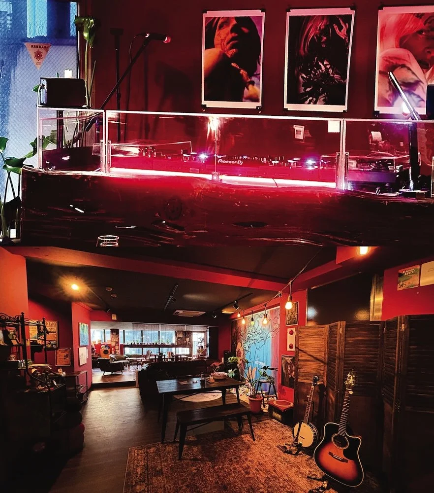 Interior of a cozy, stylish cafe or lounge with red walls and artwork, including three large framed portraits, a music setup with turntables and guitars, and warm lighting.
