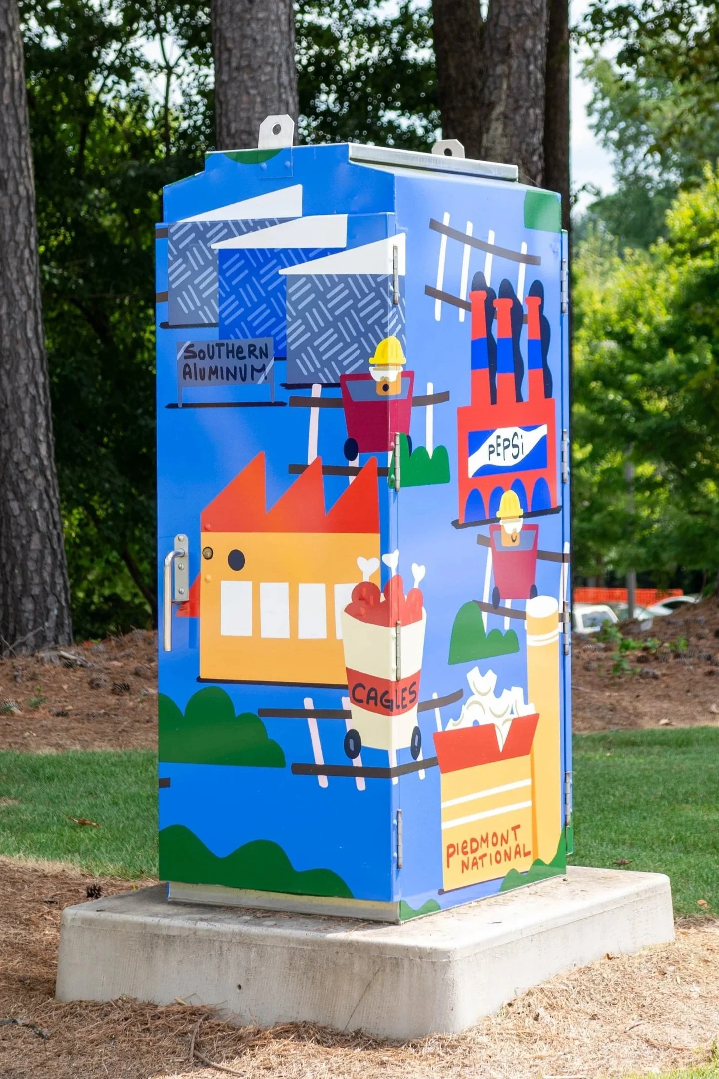 Colorful outdoor utility box painted with a vibrant mural depicting a mountain, factory, recycling bin labeled "CAGLES," and various industrial and environmental elements, with a background of green trees and a bright sky.