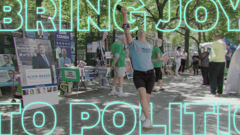 A man with a baseball cap and shirt, holding a wine glass up high, dancing at a political event with booths and banners in a park.