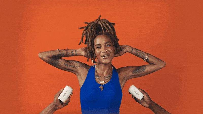 A woman with dreadlocks and tattoos is smiling with her hands behind her head, standing against an orange background. Two hands are holding white bottles labeled 'salve' near her shoulders.