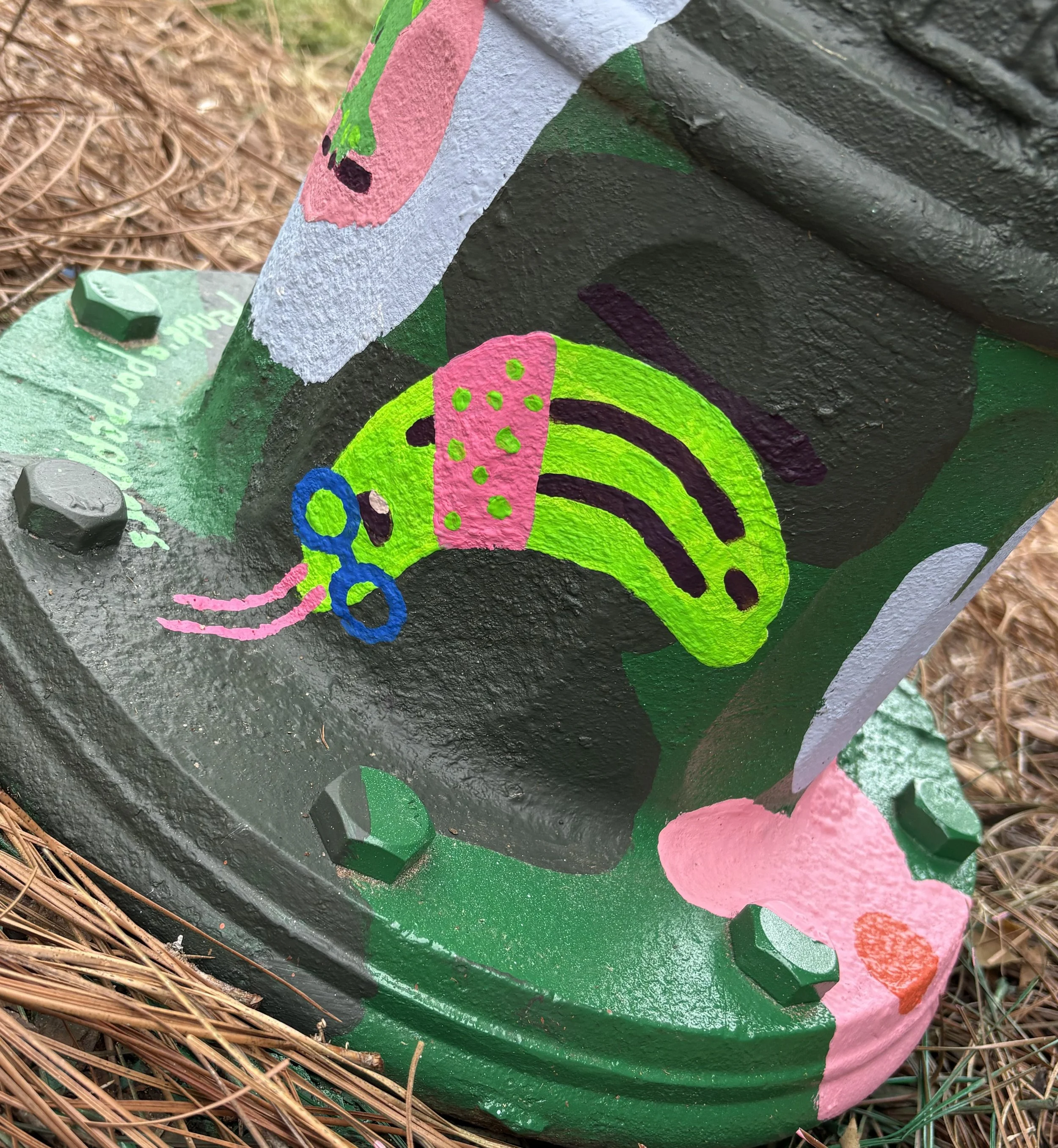 Painted outdoor utility box featuring a colorful caterpillar with pink, green, and blue sections, surrounded by pink strawberry shapes against a green and dark background, situated on a bed of pine needles.