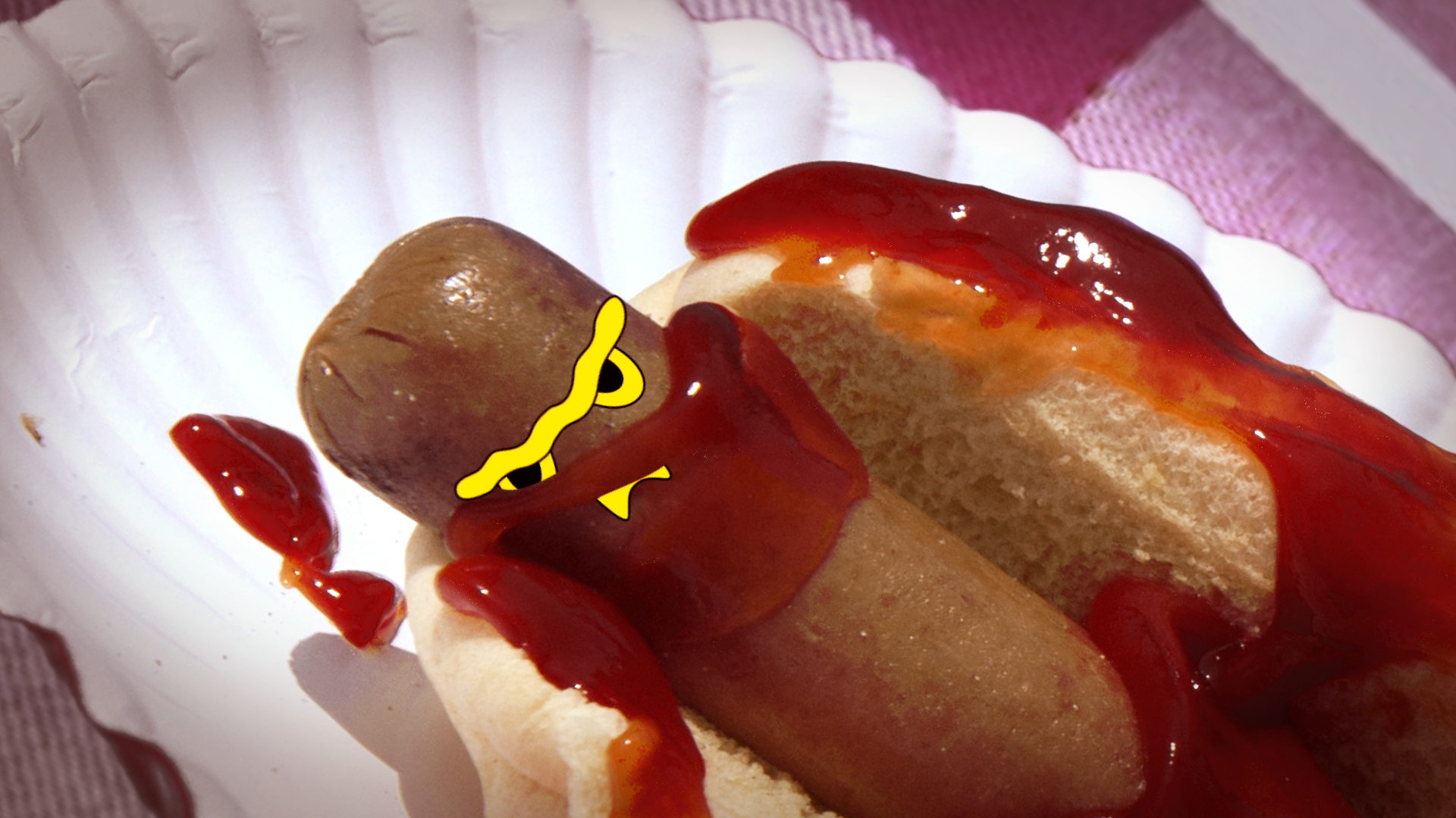 Close-up of a hot dog with ketchup on a white paper plate, with the hot dog partially covered by a bun, and a drawn cartoon face with yellow lines and a sinister expression added to the hot dog.