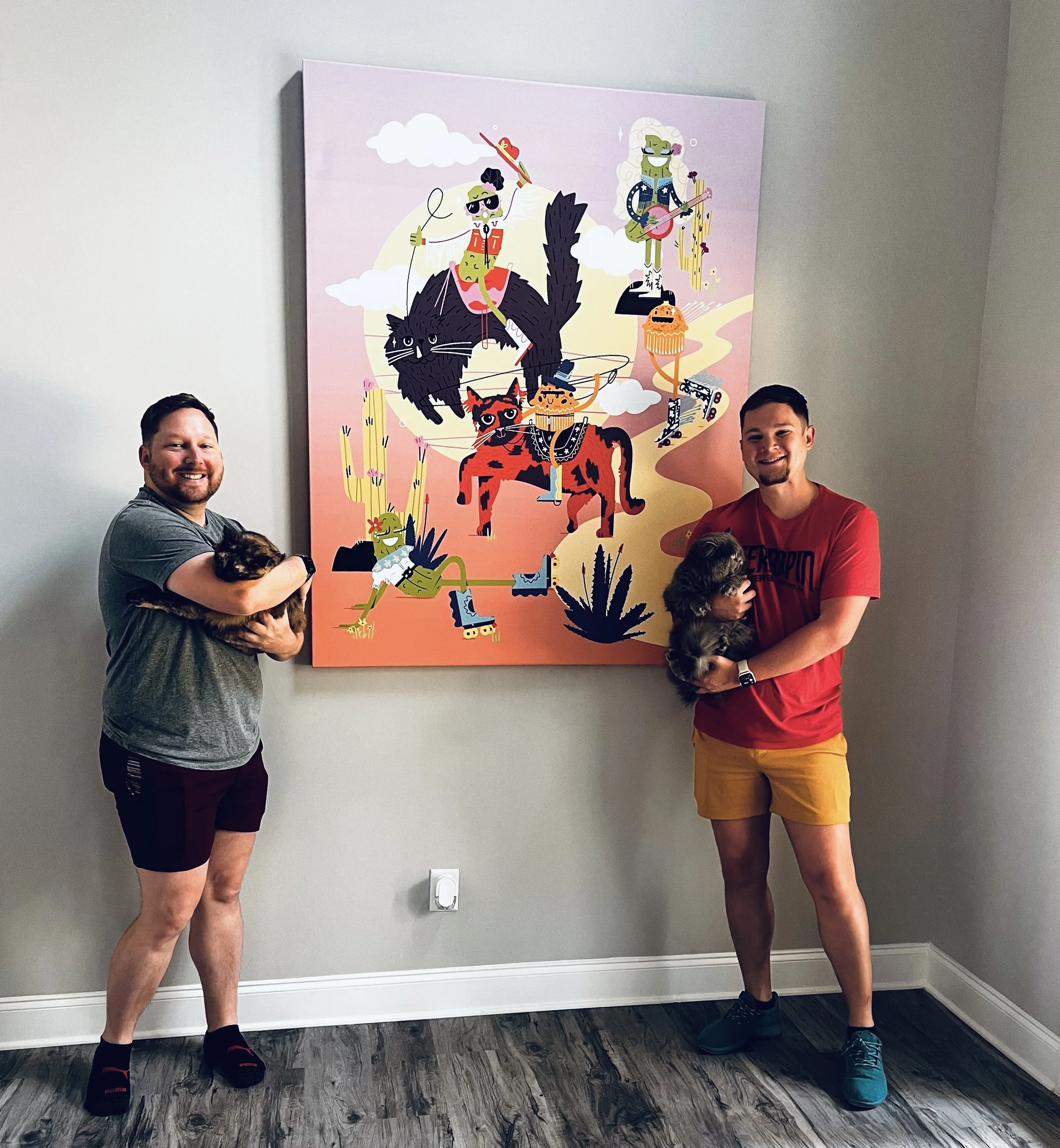 Two men smiling and holding cats in front of a colorful illustration on the wall. One man is wearing a gray t-shirt and shorts, and the other is wearing a red t-shirt and yellow shorts.