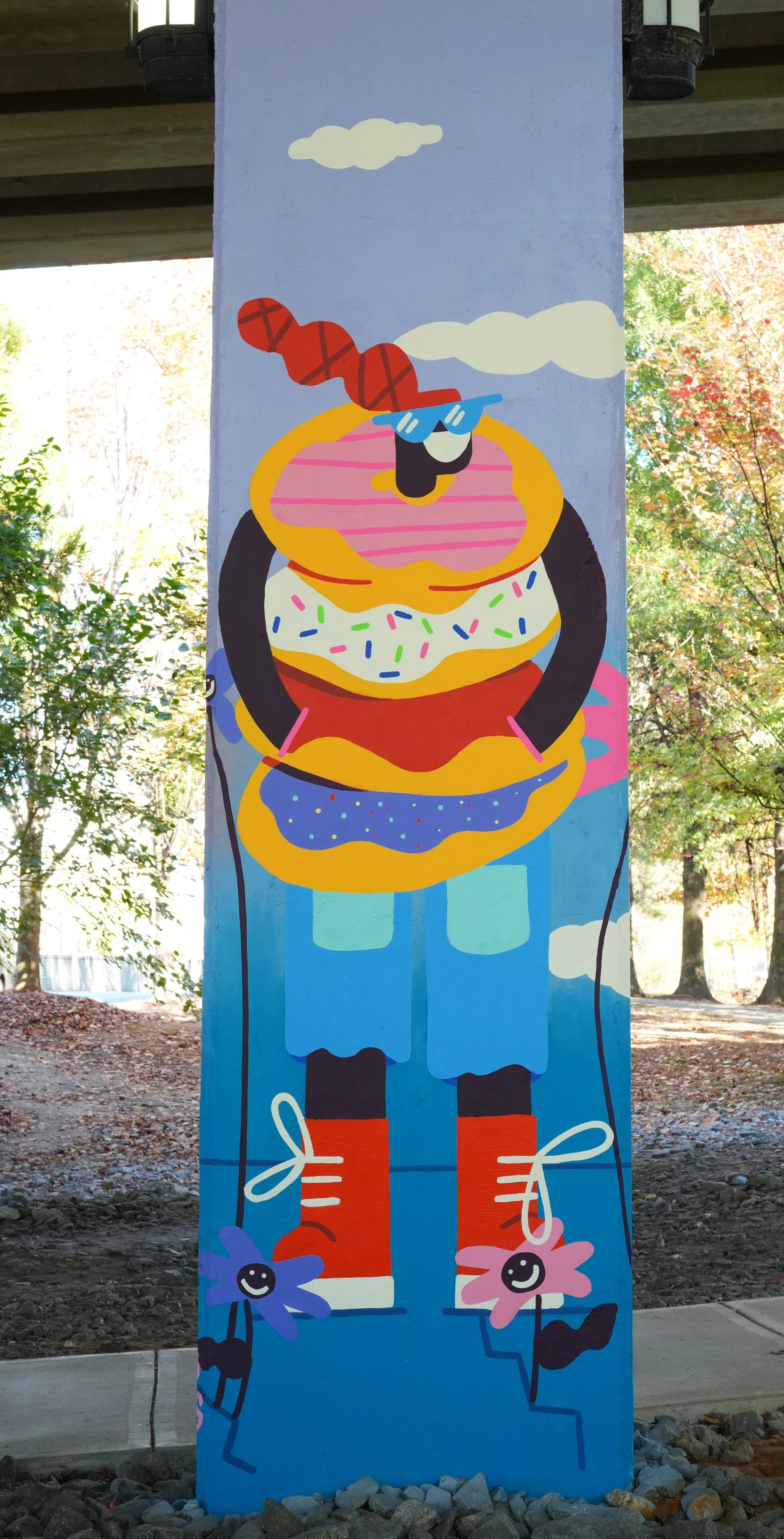 Colorful mural of a person with oversized food items as a body, wearing glasses and orange shoes, holding flower-shaped balloons, with trees and sky in the background.