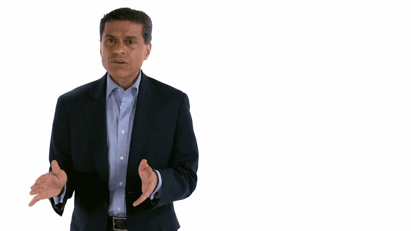 A man in a dark navy blazer and light blue shirt standing against a white background, gesturing with his hands as he speaks.