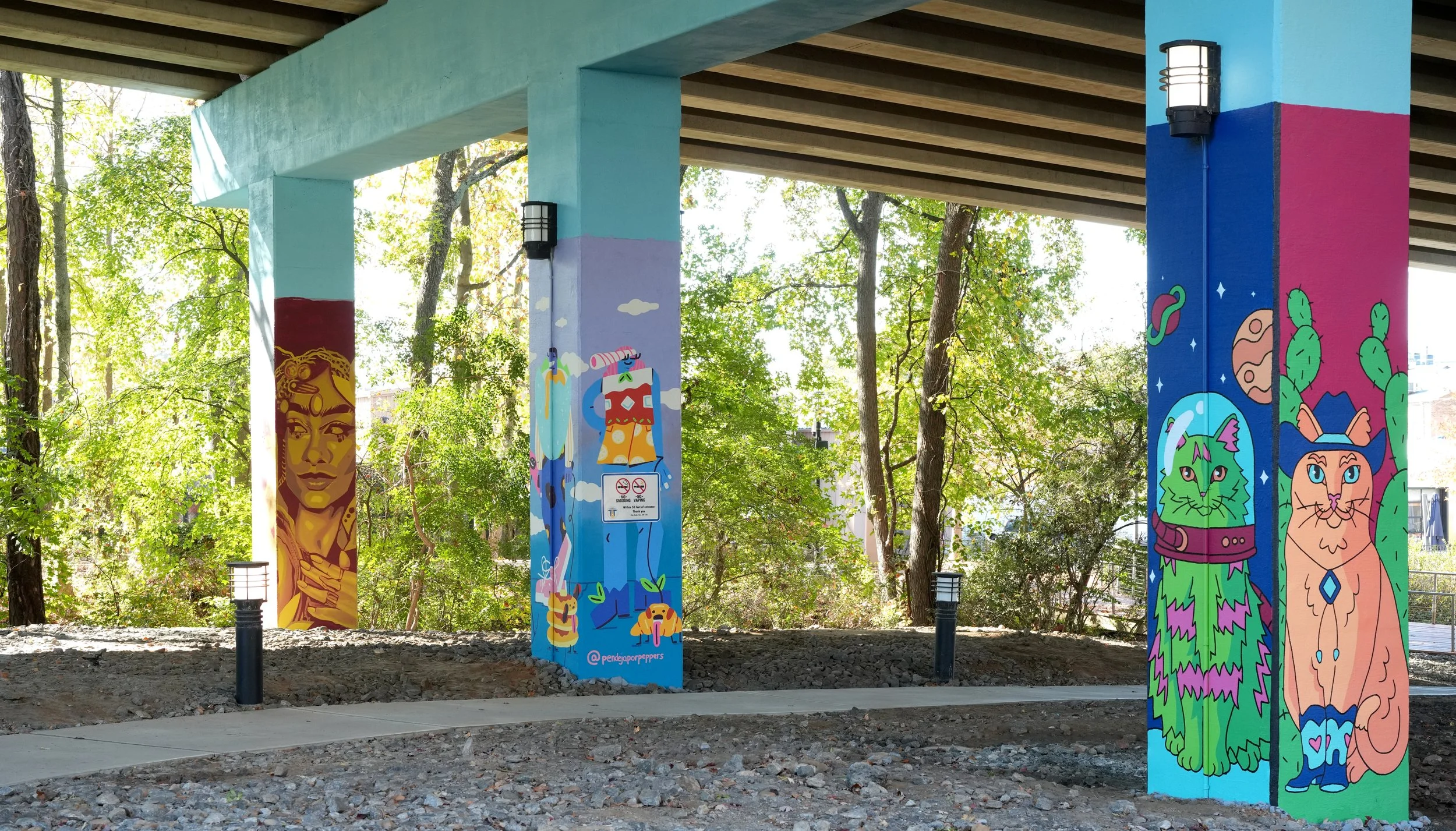 Colorful mural art on the pillars of a bridge depicting a woman with long hair and a crown, a cartoon llama with cacti, a space-themed scene with a green feline and a pink feline, and cartoon planets and stars.