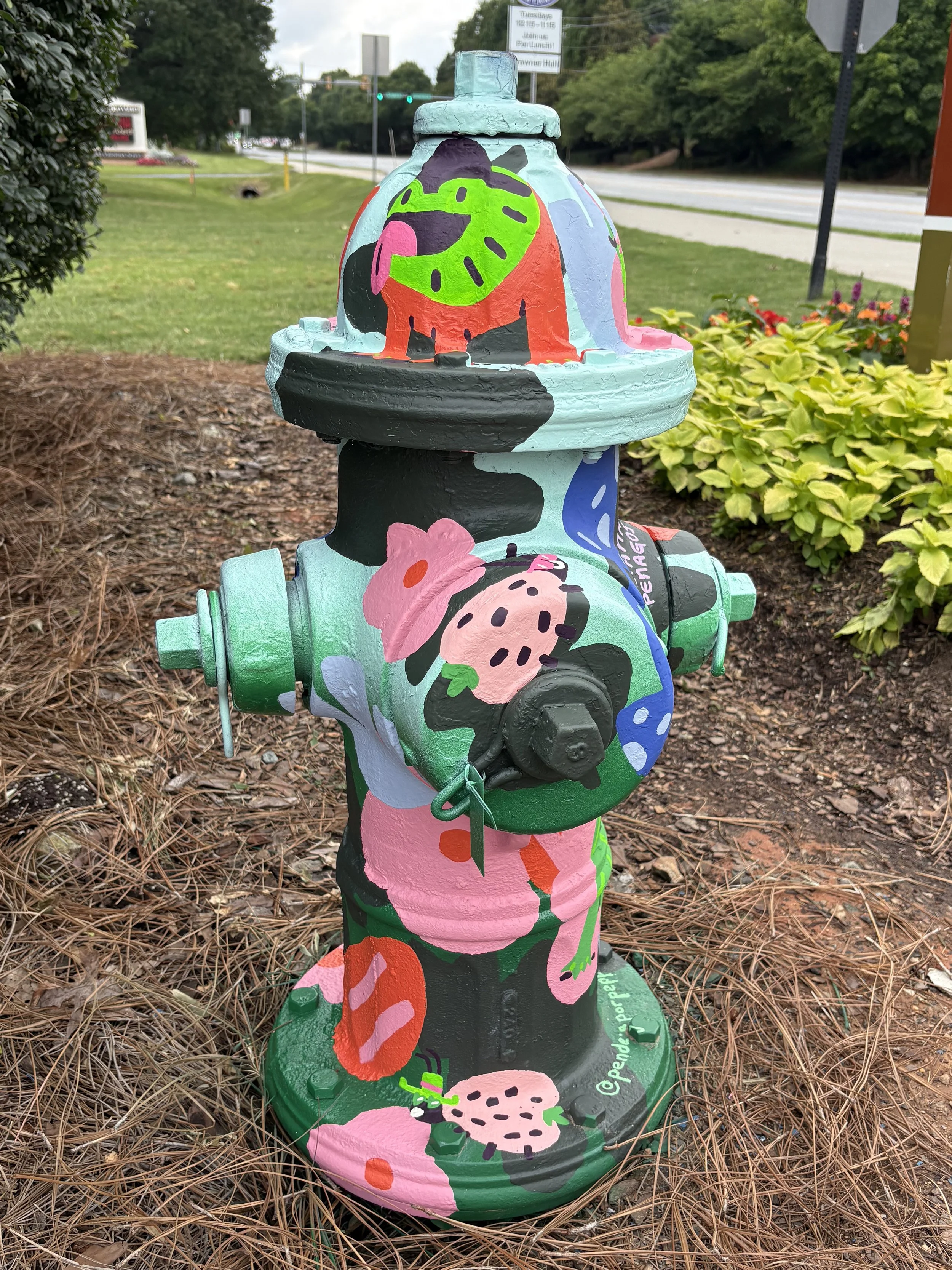 Colorfully painted fire hydrant with floral and leaf patterns in pink, orange, green, black, and blue, situated on a mulch bed with bushes and a street background.