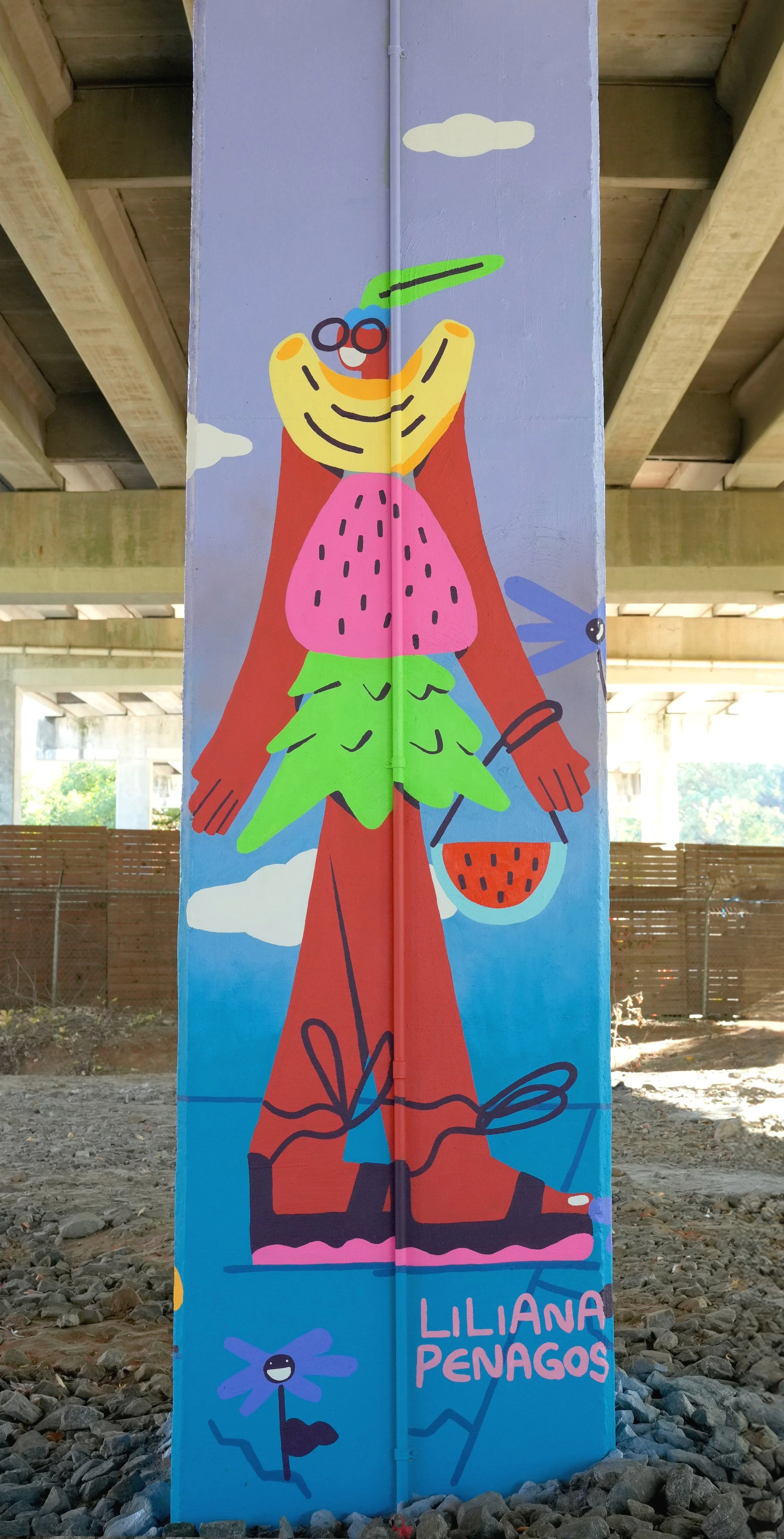 Colorful mural of a smiling person dressed as a butterfly with green wings, pink body, red pants, and holding an orange bag, with clouds and dragonflies in the sky, signed by Liliana Penagos.