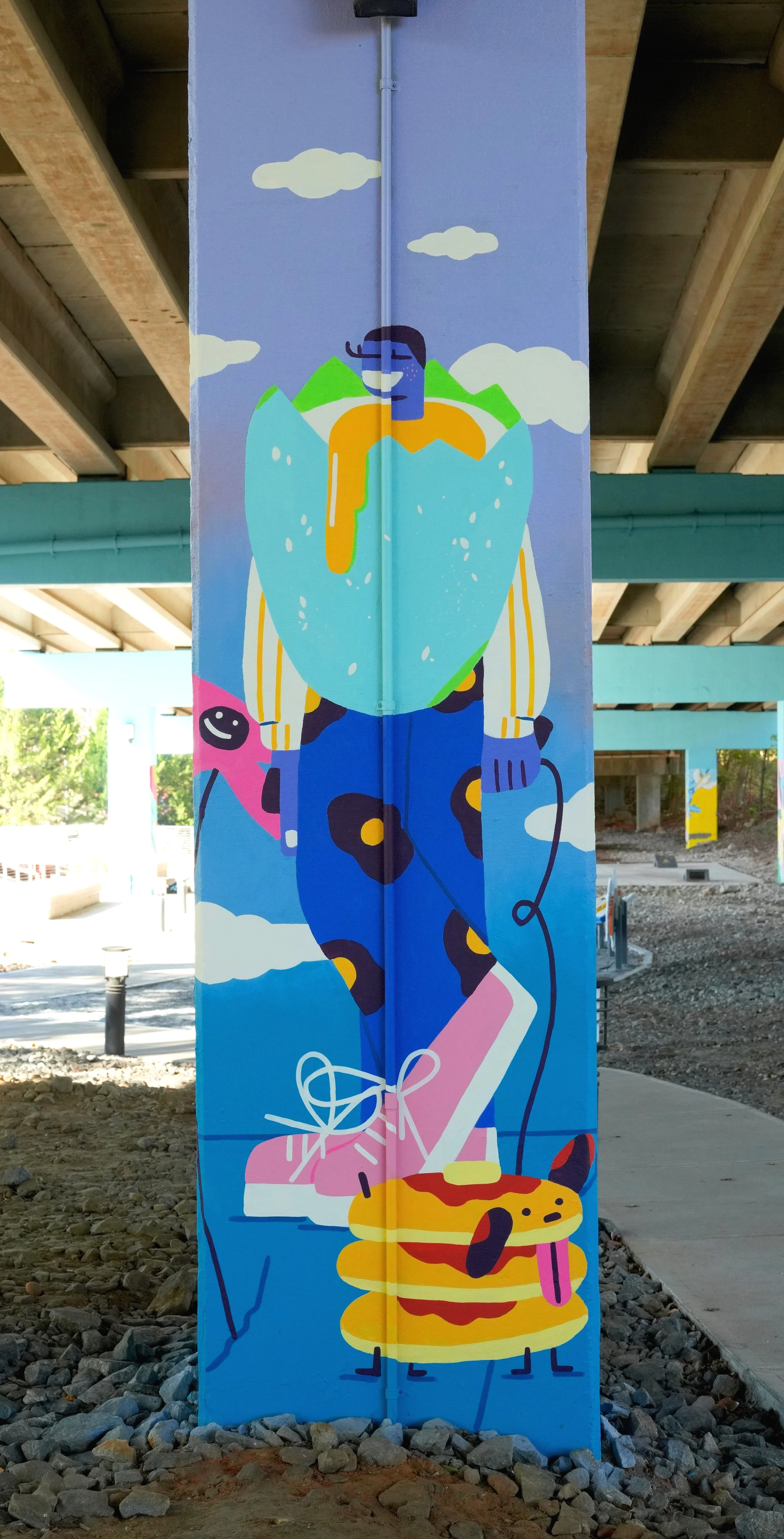 Colorful mural of a person with a large blue and yellow cloud, holding a pink device connected to a cartoon dog made of pancakes with syrup and a tongue, under a blue sky with clouds, in an urban setting under a bridge.