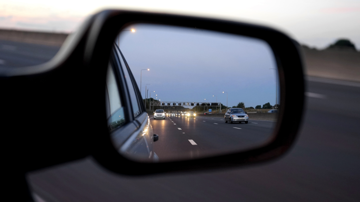 Vue d'une autoroute depuis le rétroviseur d'une voiture, avec plusieurs véhicules en arrière-plan sous un ciel crépusculaire.