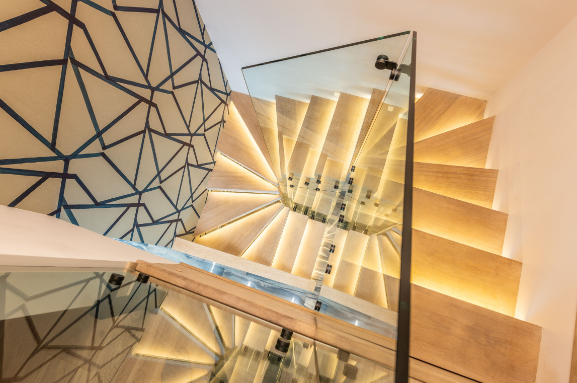 Escalier en colimaçon en bois avec balustrade en verre illuminée, mur avec motif géométrique.