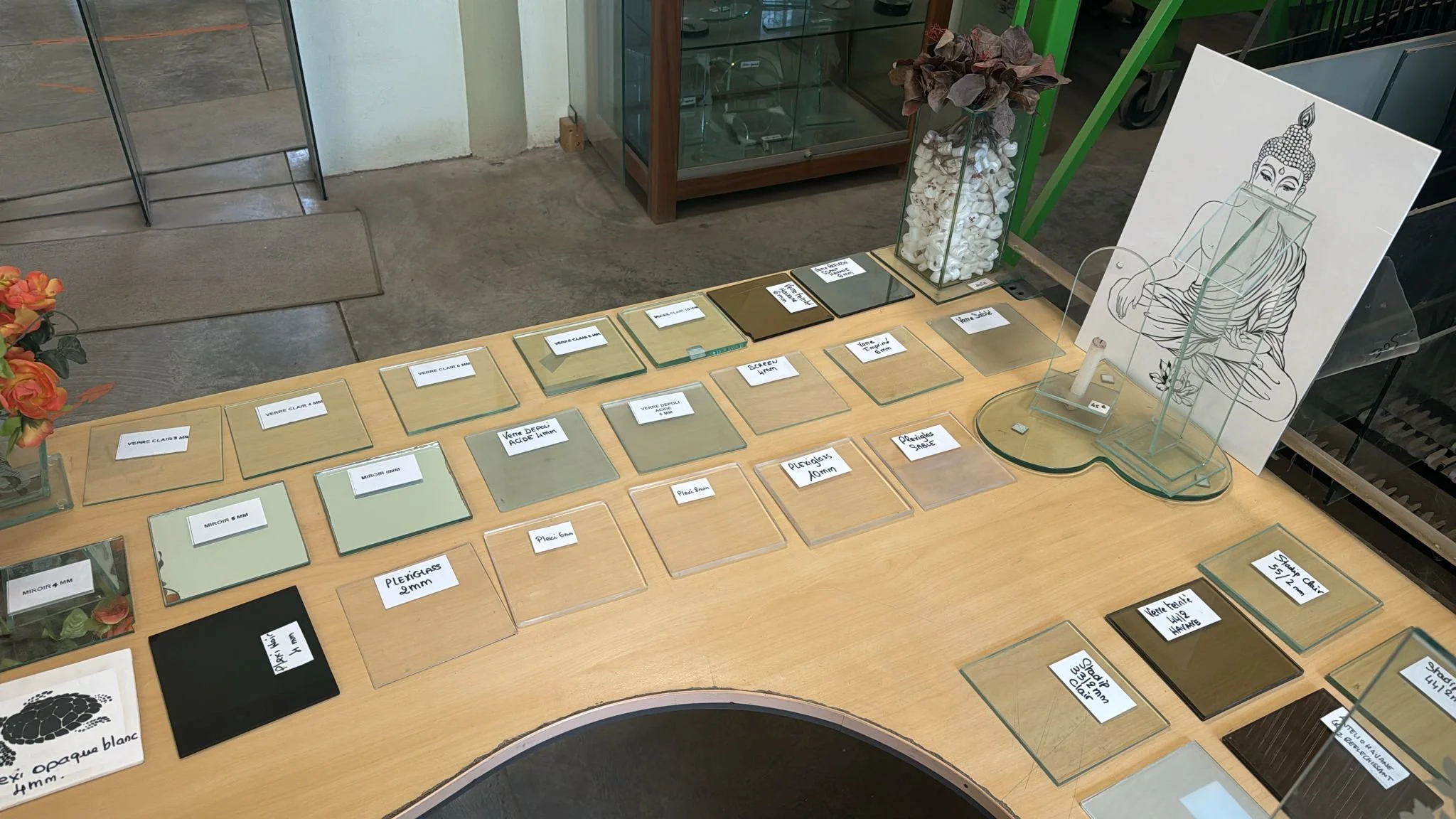 Des échantillons de différents types de verre, chacun étiqueté avec sa description, sont disposés sur une table en bois. Il y a également des décorations florales et un dessin de Bouddha en arrière-plan. Les étiquettes indiquent des informations sur l'épaisseur et le type de verre, comme le plexiglas et le verre coloré.