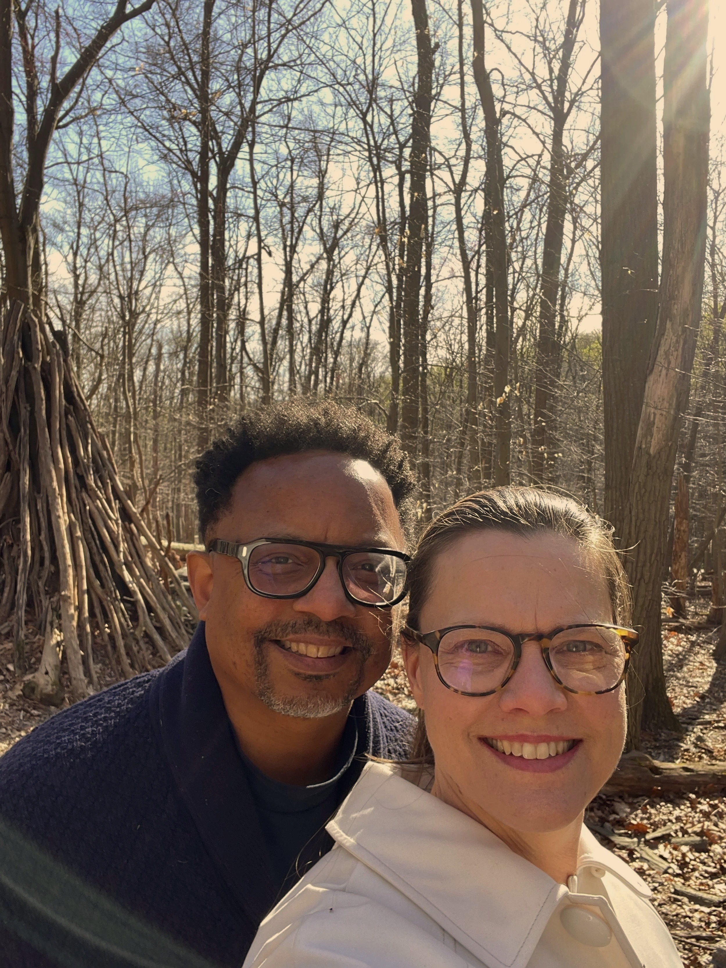 A smiling man with curly hair and glasses, and a smiling woman with glasses, taking a selfie outdoors in a wooded area on a sunny day.
