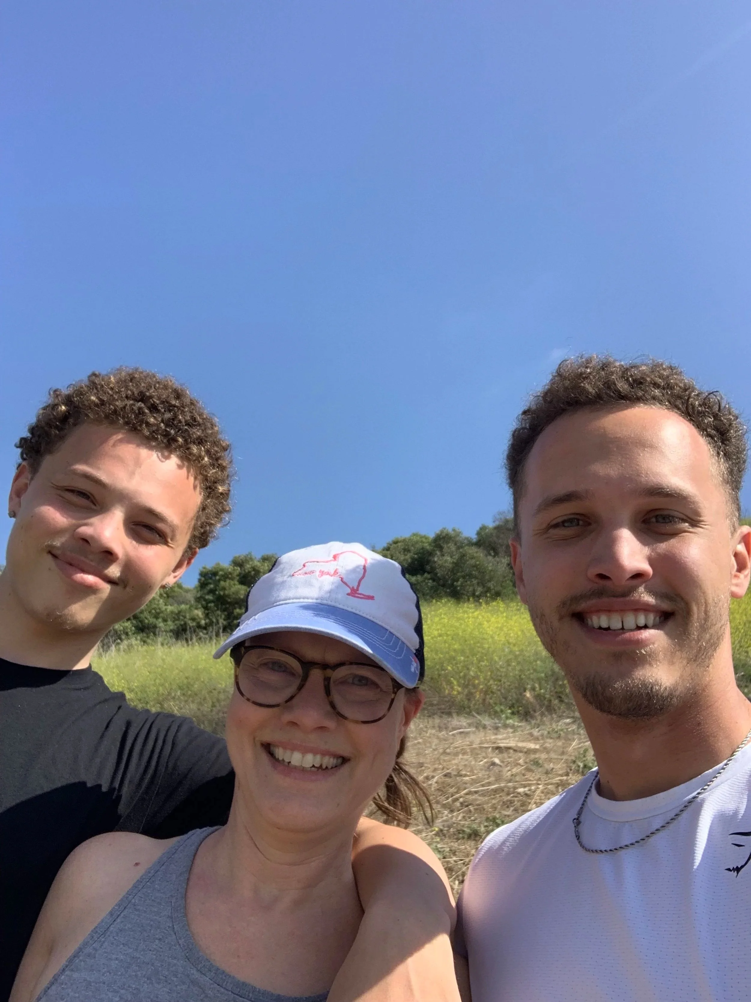 A group of four people taking a selfie outdoors on a sunny day, with green trees and yellow flowers in the background and a clear blue sky.