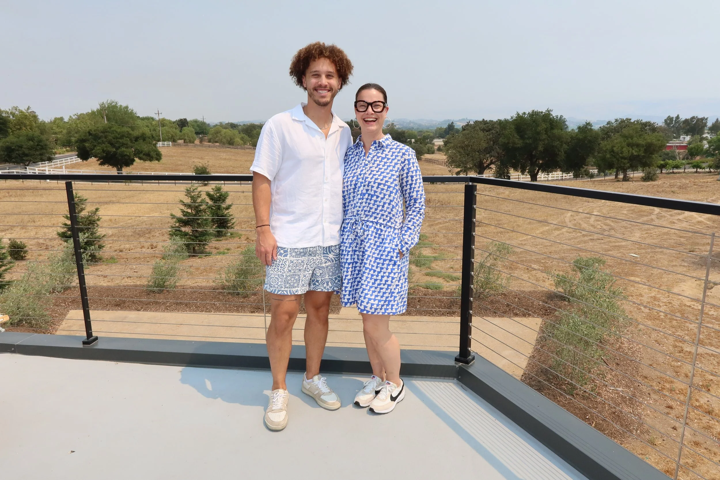 Deb Valentine standing outdoors on a deck with one of her adult sons, Josh, both smiling, with rolling hills and trees in the background under a clear sky.