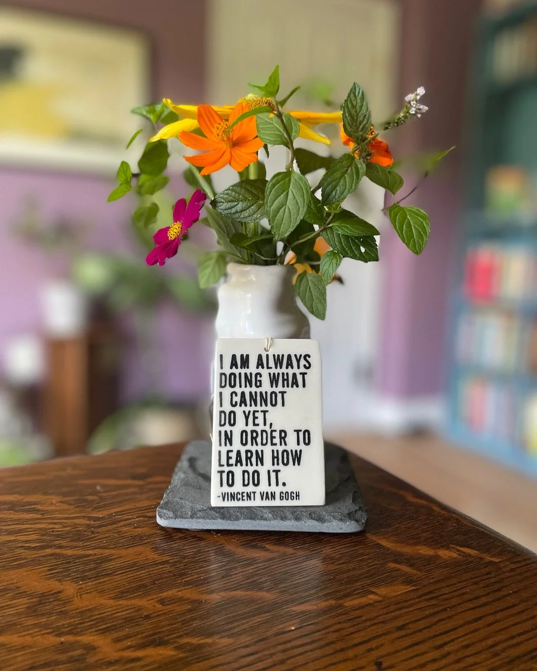 A white ceramic vase on a wooden table holding colorful flowers and greenery, with a tag that reads, “I am always doing what I cannot do yet, in order to learn how to do it,” by Vincent van Gogh.