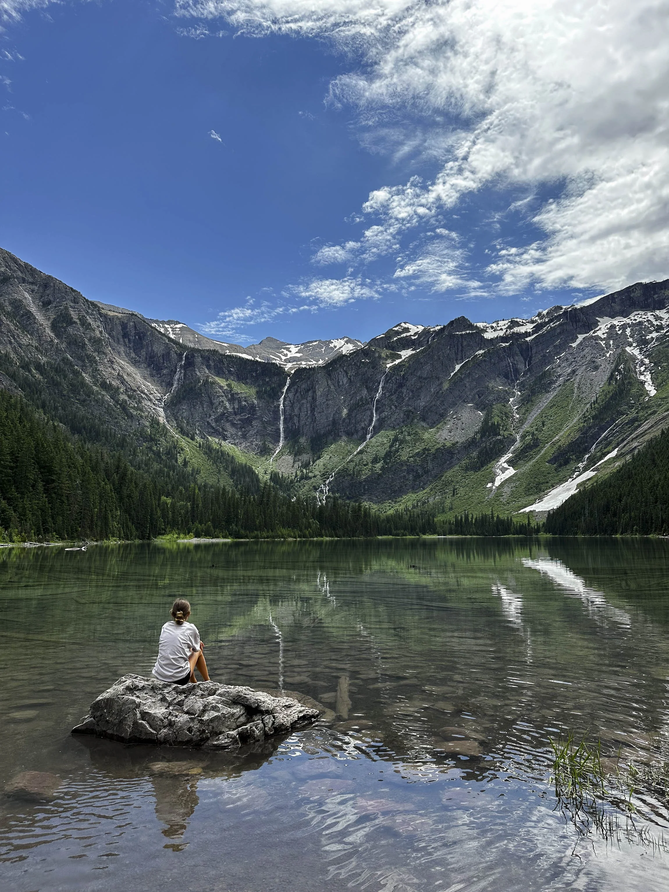 A person sitting on a large rock at the edge of a calm lake, surrounded by green trees, with mountains in the background and a partly cloudy sky.