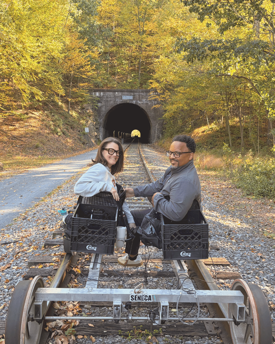 A man and a woman smiling and riding a rail bike on a track leading into a tunnel surrounded by autumn foliage.